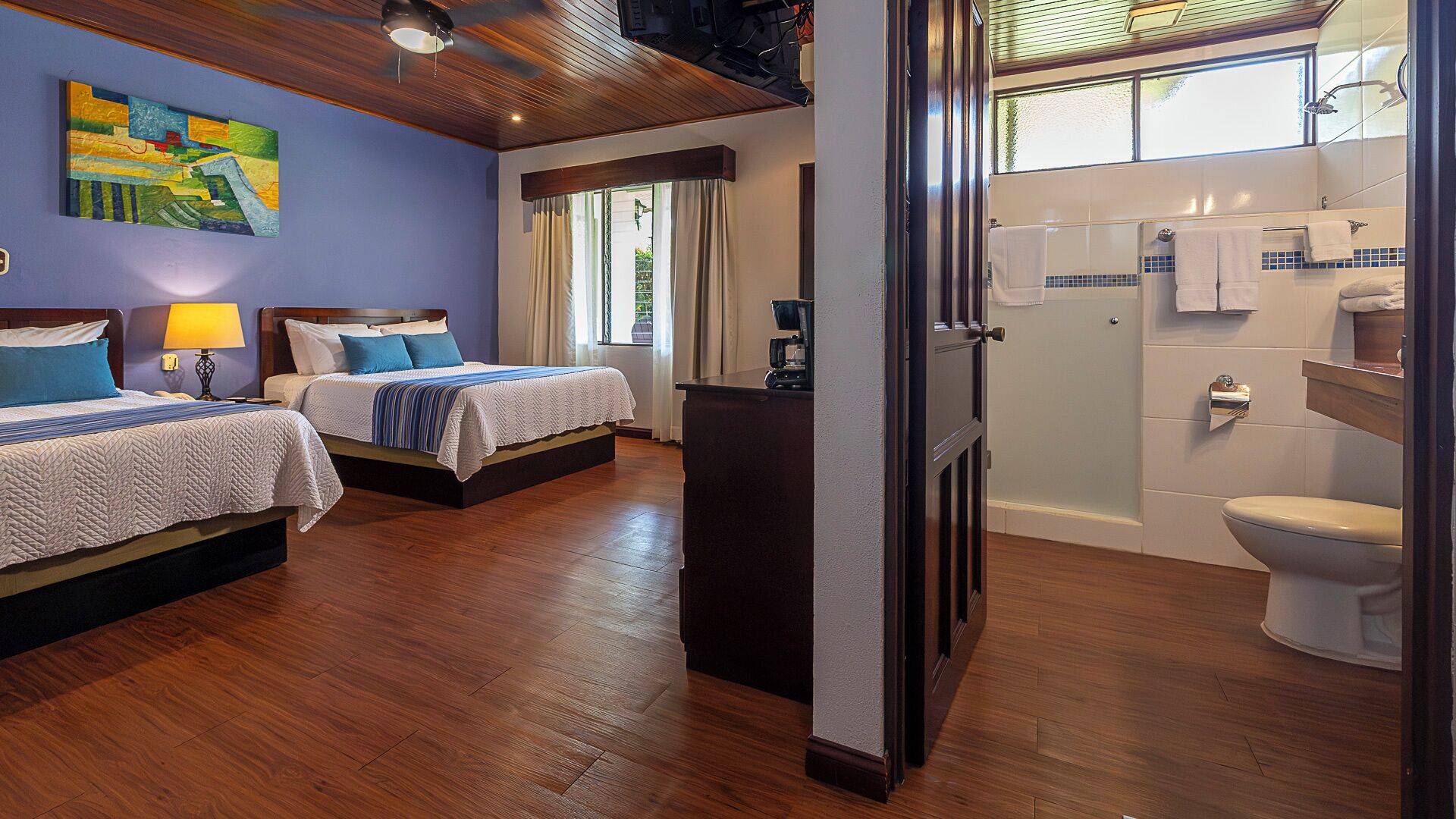 A hotel room with two beds, a painting on the wall, a window with curtains, and a bathroom with a toilet and sink.
