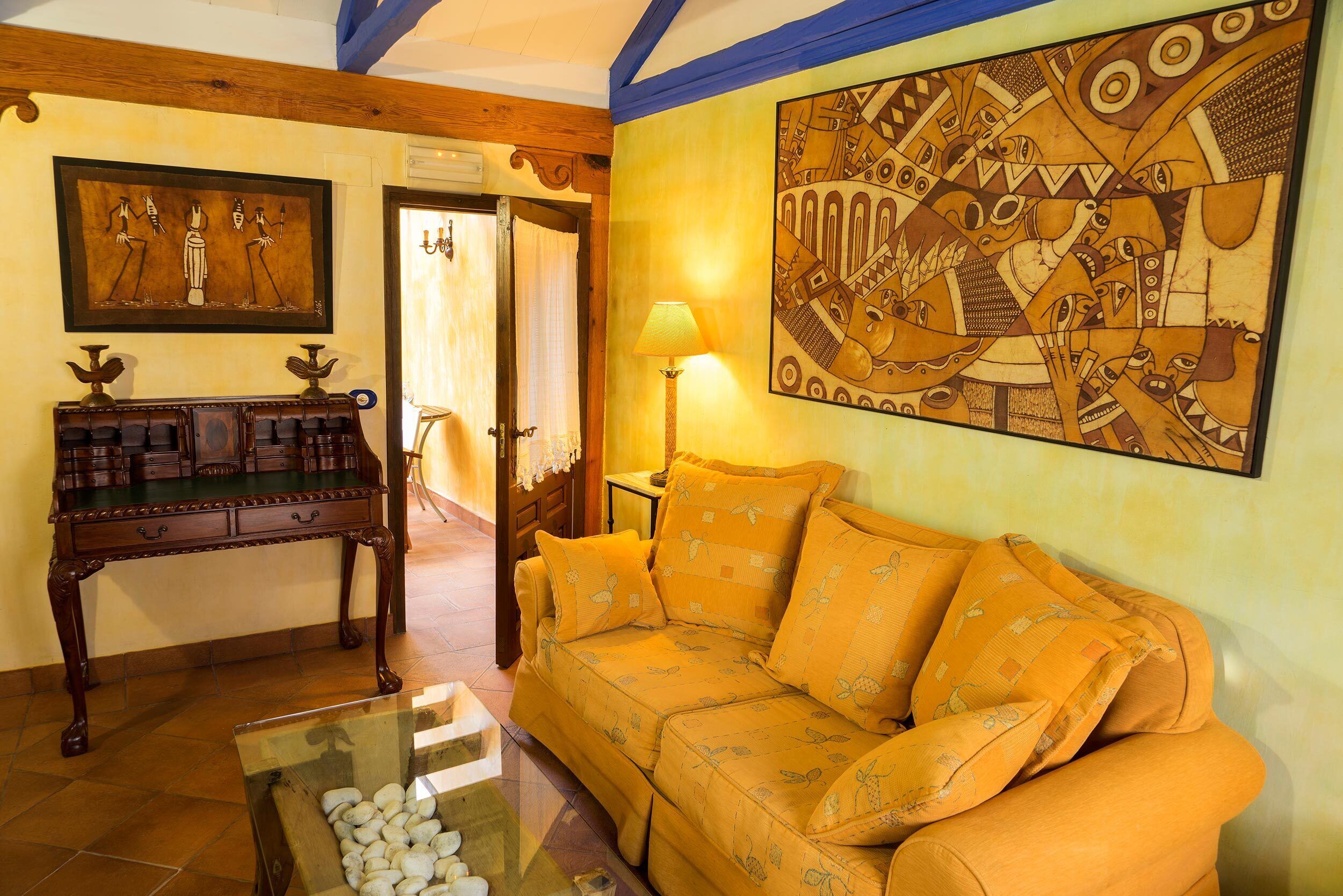 A room with a wooden coffee table, a sofa with patterned cushions, a wooden side table, and a wall with a large abstract painting.