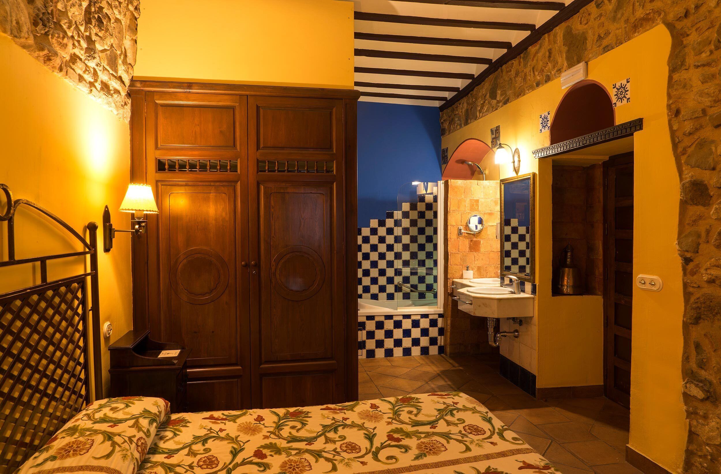 Een slaapkamer met een bed, een houten garderobe, een nachtkastje en een badkamer met een geruit vloertje, een wastafel en een spiegel.