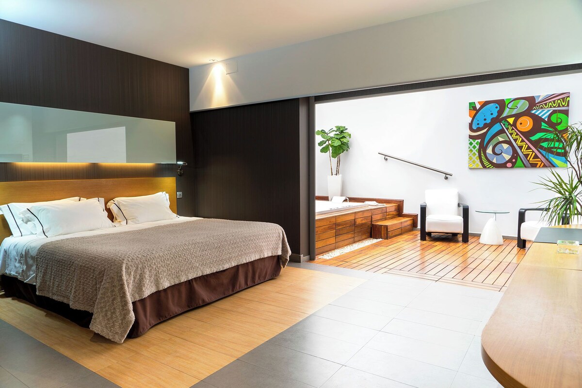 Un dormitorio moderno con una cama grande, cabecera de madera, una mesita de noche y un mueble auxiliar. Hay una ventana con vistas a un balcón y un cuadro abstracto colorido en la pared.