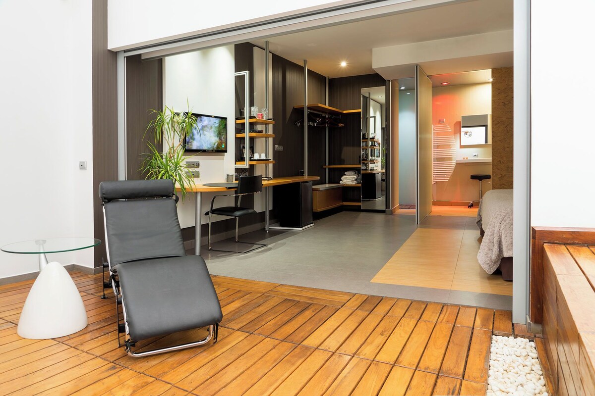 Una habitación moderna con suelo de madera, un sillón negro, una mesa de vidrio y un televisor en la pared.