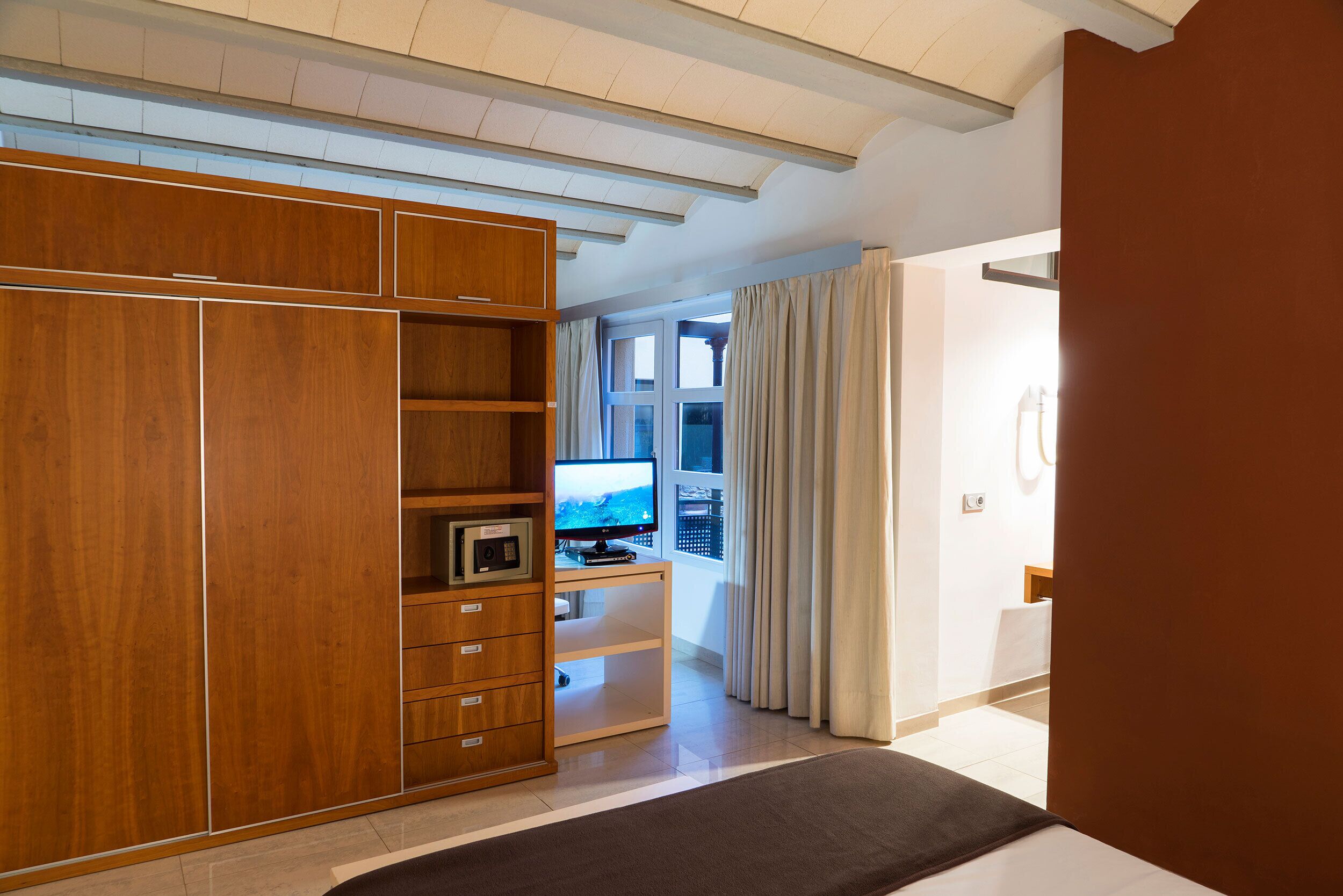 A bedroom with a large wardrobe, a bed, a desk with a computer, and a window with curtains.