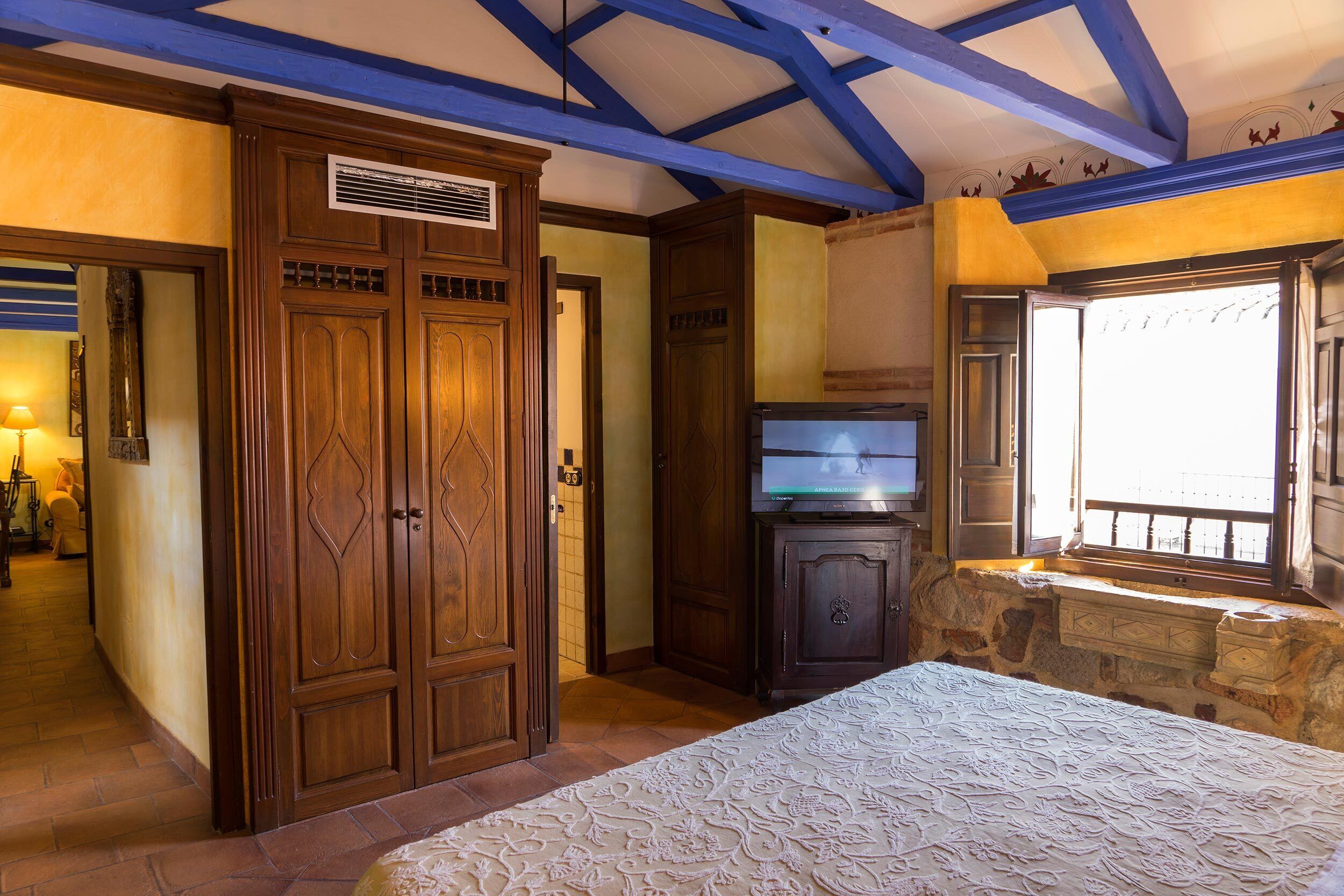 A bedroom with a bed, wooden wardrobe, television, stone wall, and a window with a view.