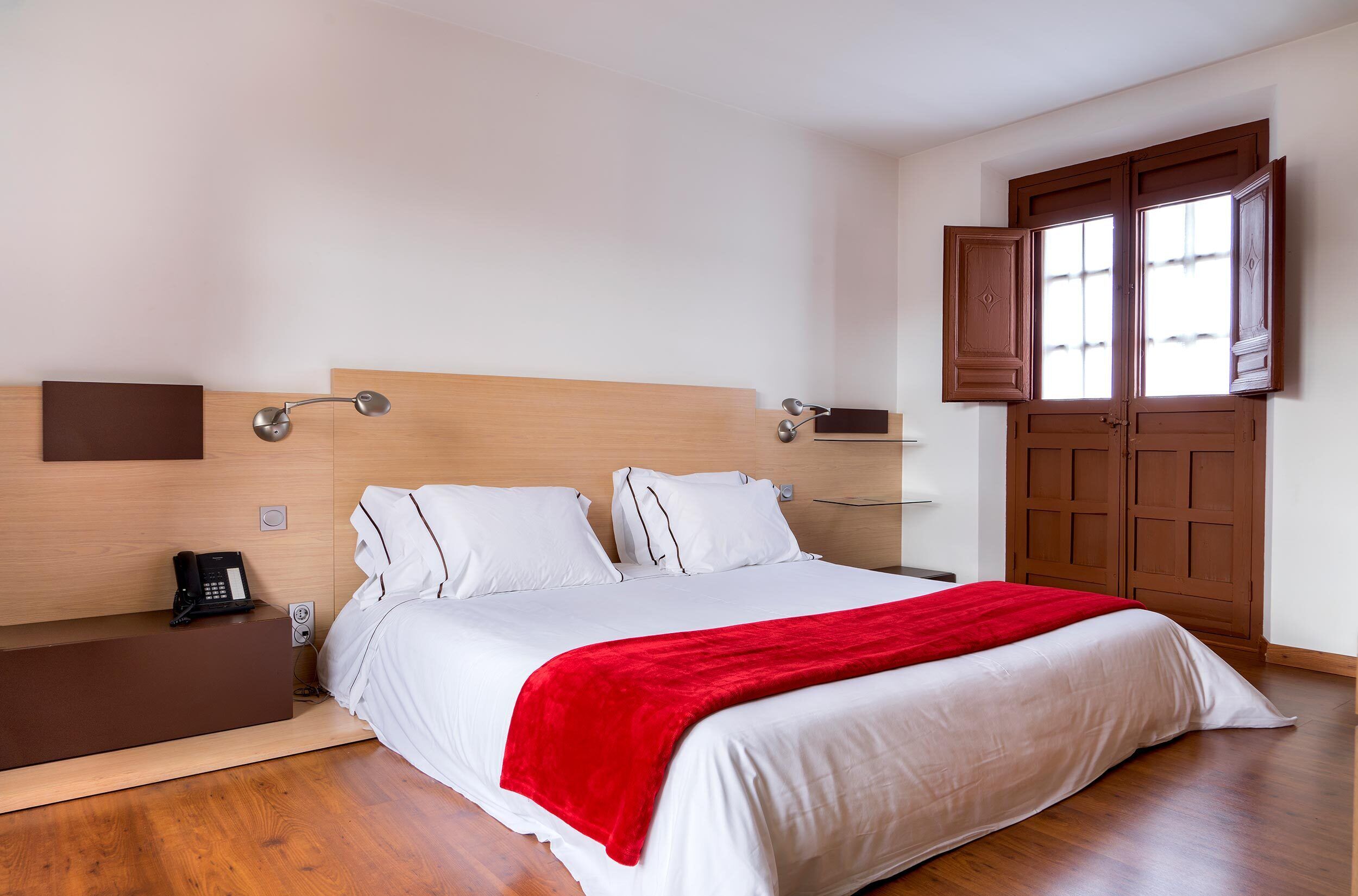 A neatly made bed with white linens and a red throw blanket, a bedside table with a phone, and a wooden headboard.