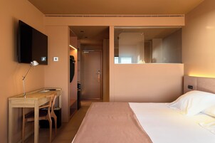 A hotel room with a bed, a desk with a chair, a TV mounted on the wall, and a door leading to another room.