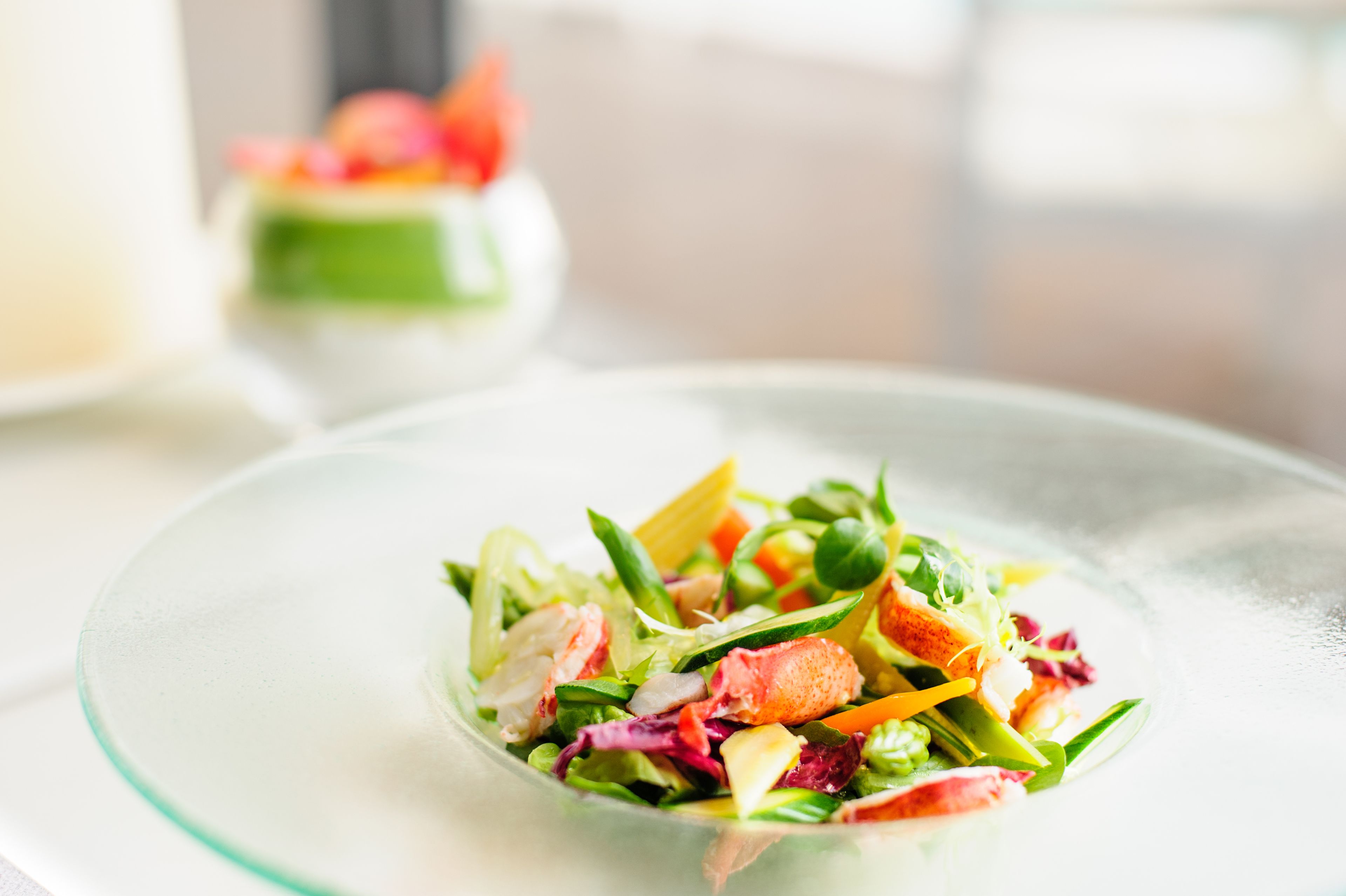 A clear glass bowl of mixed salad with various vegetables and a piece of salmon on a white plate.