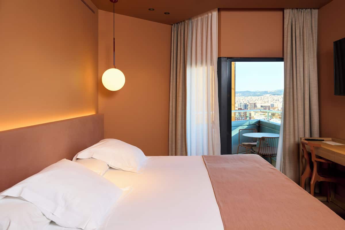 Quarto de hotel com uma cama grande, uma mesa e vista para o horizonte urbano através de uma porta deslizante.