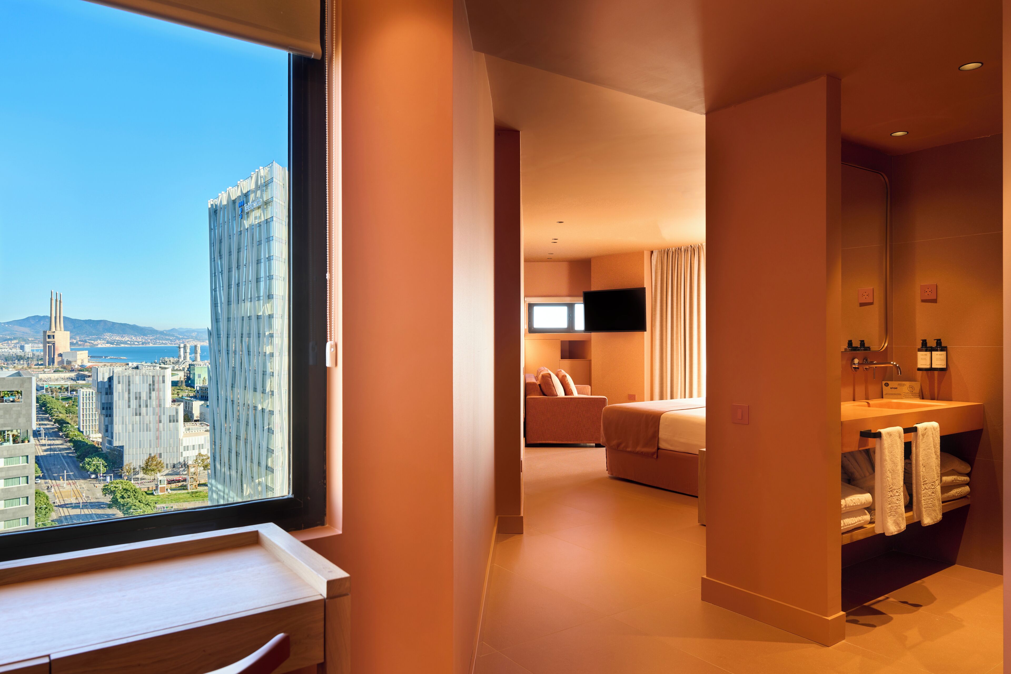 Habitación de hotel moderna con vistas a la ciudad, una ventana grande, cama, televisor y baño con lavamanos y toallero.