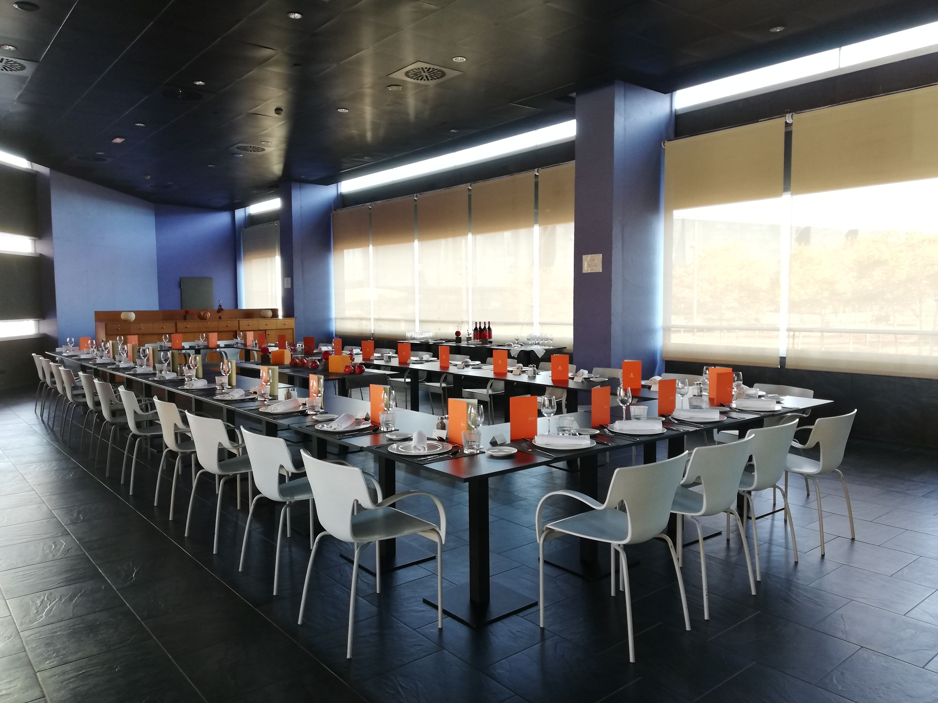 Un amplio comedor con mesas largas dispuestas para varios comensales, sillas blancas y sujetadores de menú naranja.