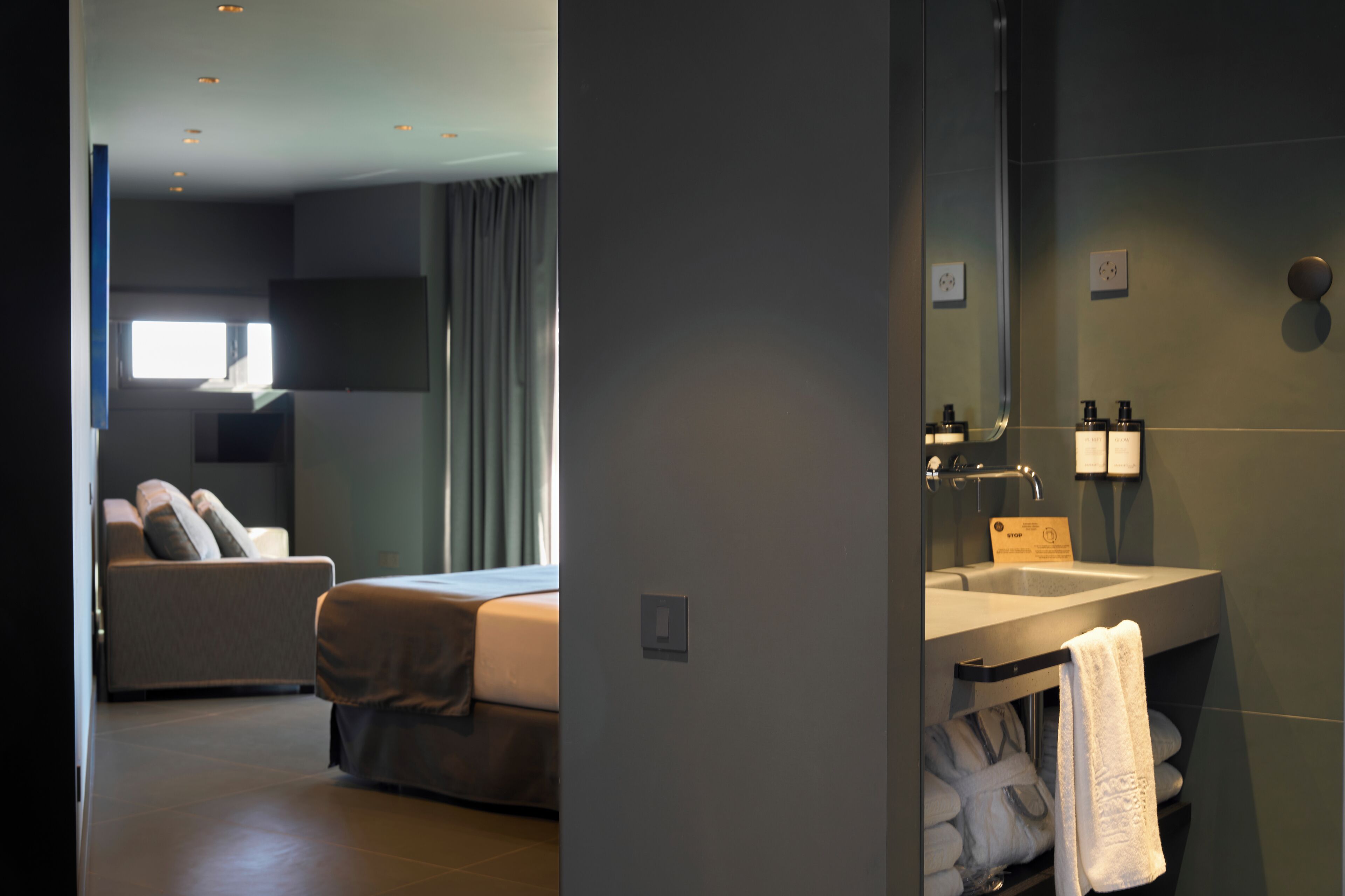 A modern hotel room with a bed, a sofa, a large window, and a bathroom with a sink and towel rack.