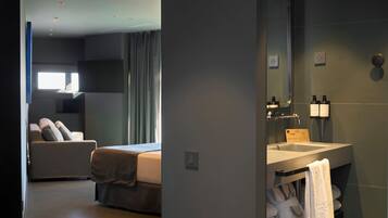 A modern hotel room with a bed, a sofa, a large window, and a bathroom with a sink and towel rack.