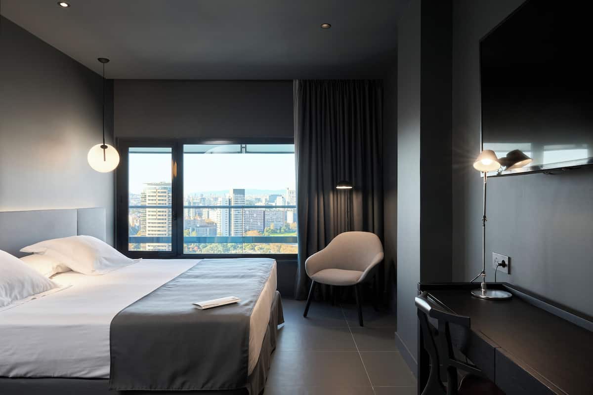 Quarto de hotel moderno com uma cama grande, uma escrivaninha, uma cadeira e vista para a cidade.