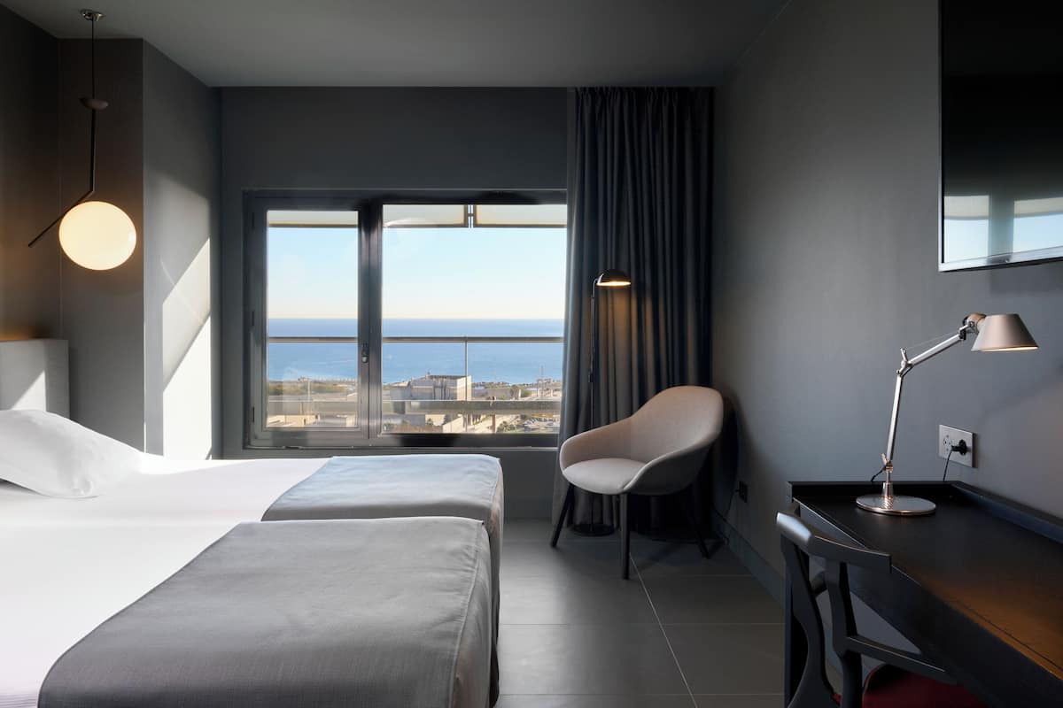 Quarto de hotel com duas camas, uma escrivaninha, uma cadeira, um abajur e uma janela grande com vista para a cidade.