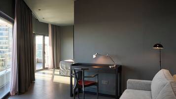 A modern hotel room with a desk, chair, and a sofa. There is a large window with curtains, a floor lamp, and a view of buildings outside.