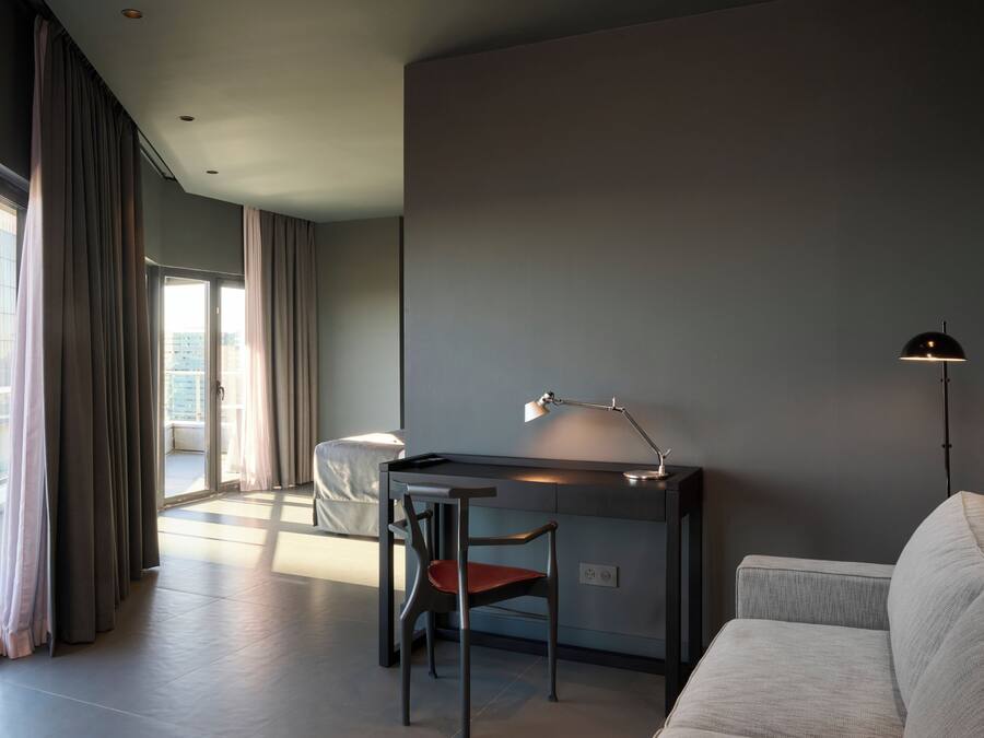 Una habitación de hotel moderna con escritorio, silla y sofá. Cuenta con un ventanal con cortinas, una lámpara de pie y vistas a edificios.