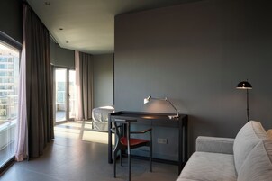 A modern hotel room with a desk, chair, and a sofa. There is a large window with curtains, a floor lamp, and a view of buildings outside.
