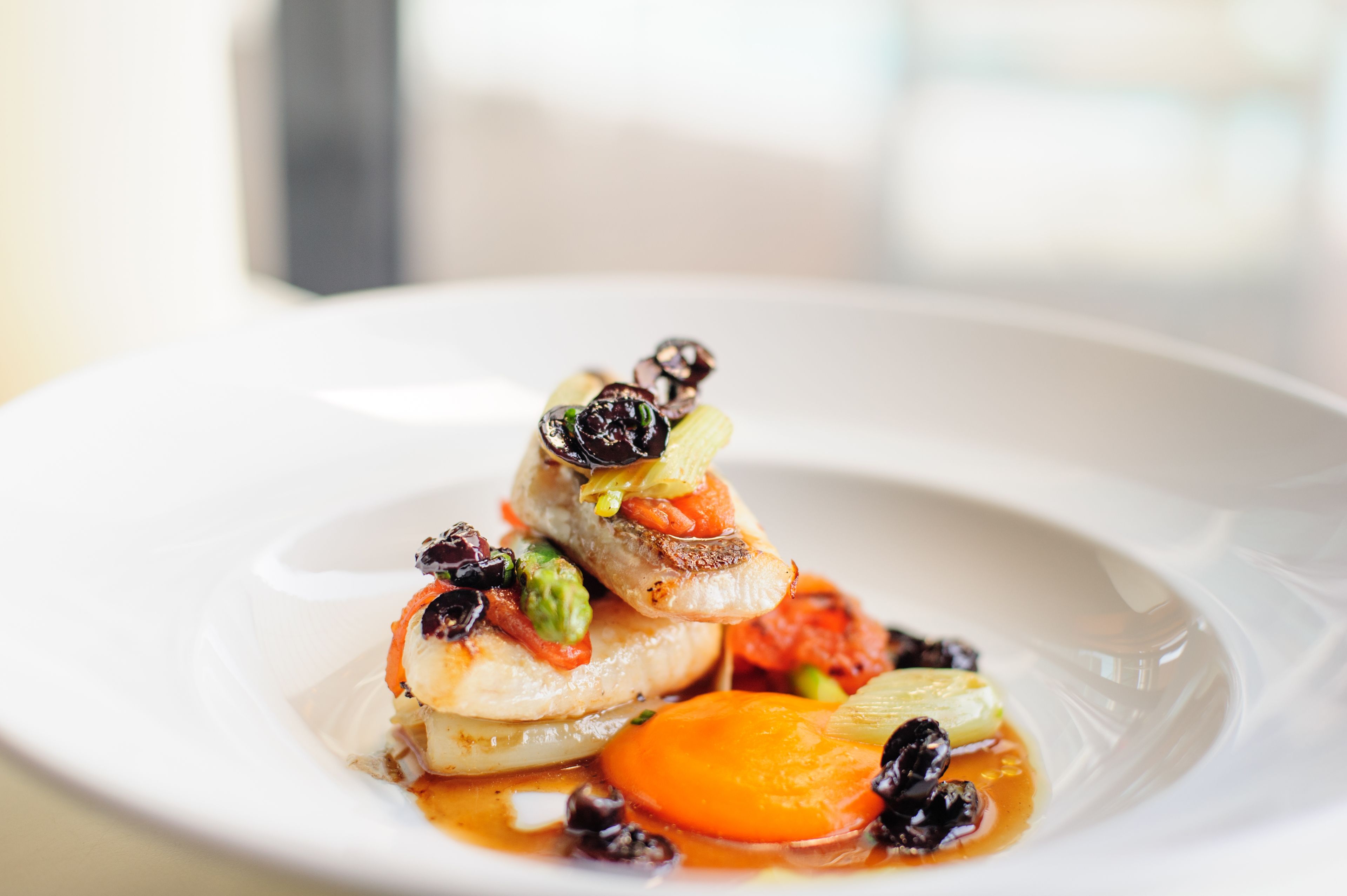 A white plate with a seafood dish, including fish, vegetables, and olives.