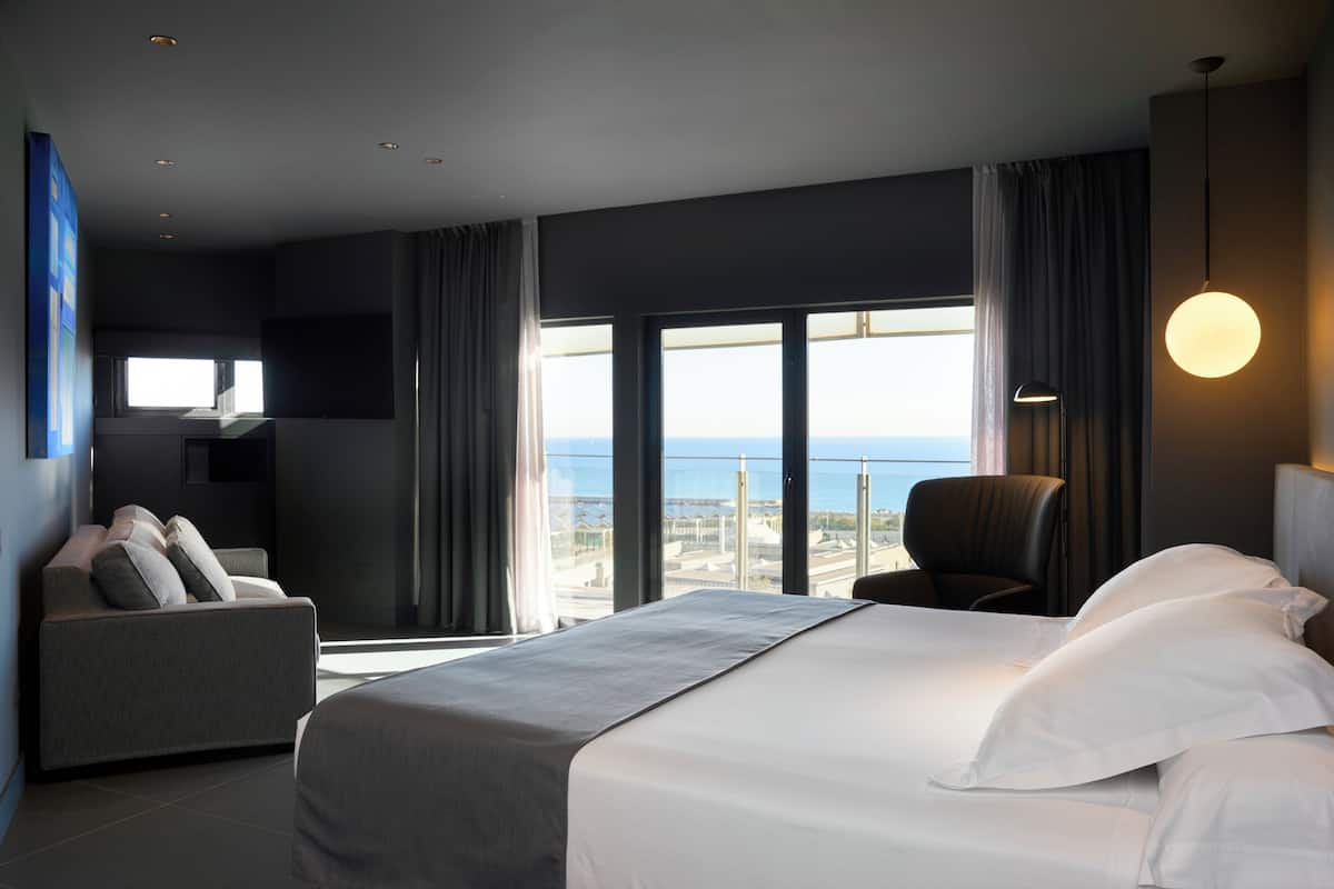 Quarto de hotel moderno com uma cama grande, um sofá, uma poltrona e vista para a cidade.