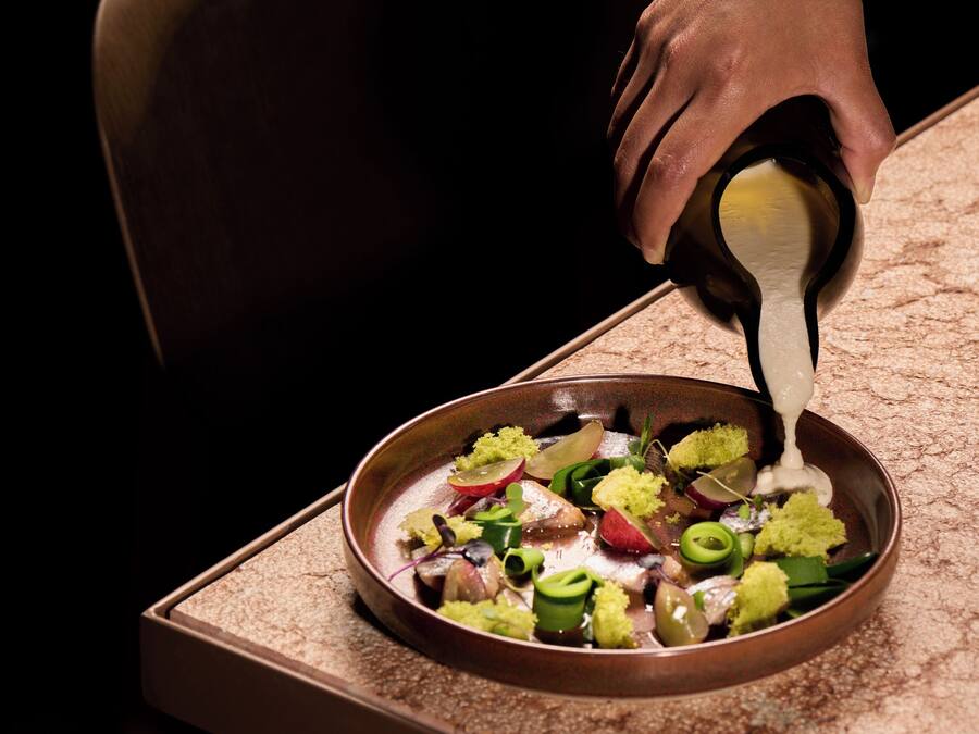 A person pouring a creamy sauce over a dish of mixed vegetables on a table.