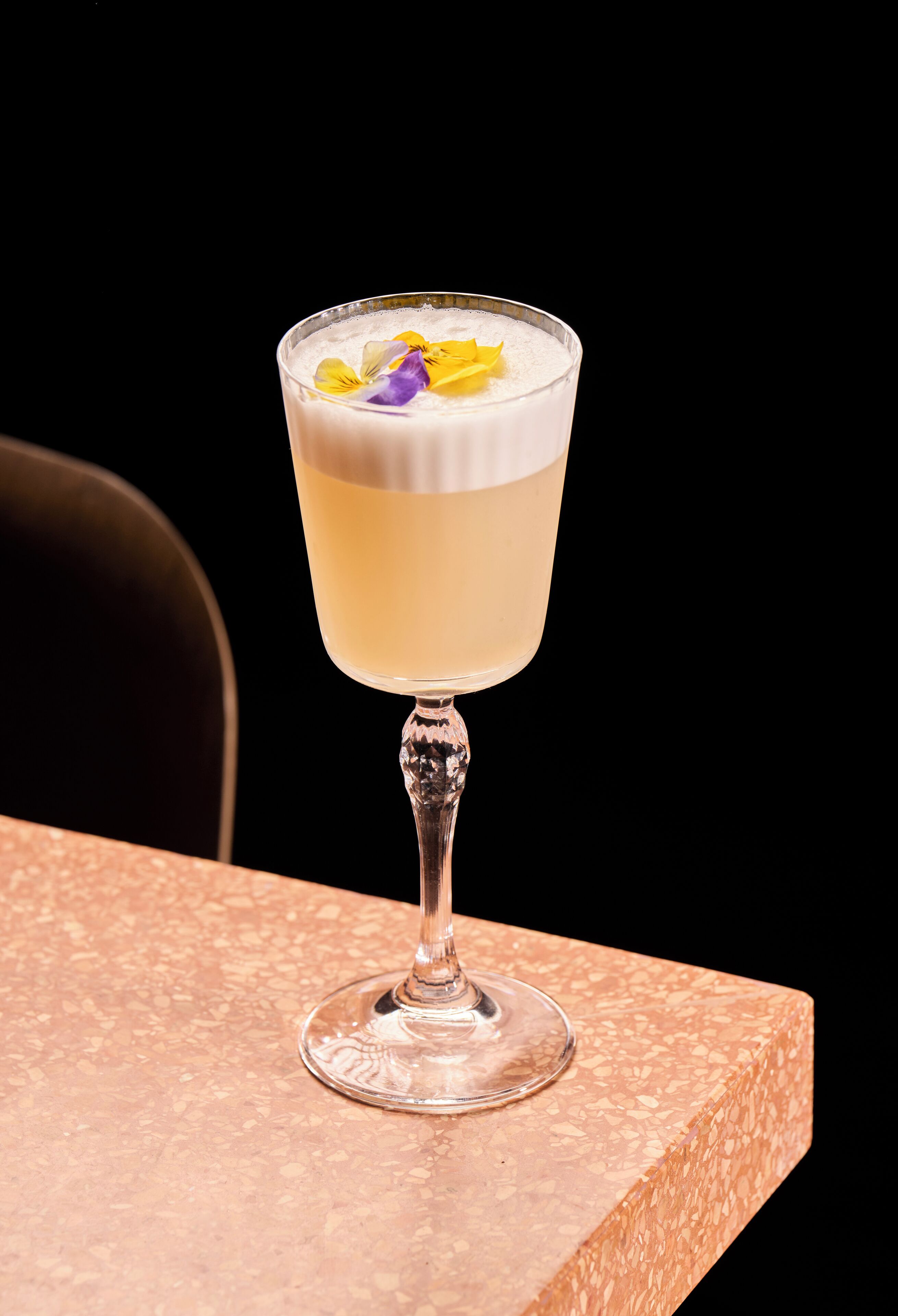A cocktail in a stemmed glass with a layered appearance and a garnish of yellow and purple flowers on a textured surface.