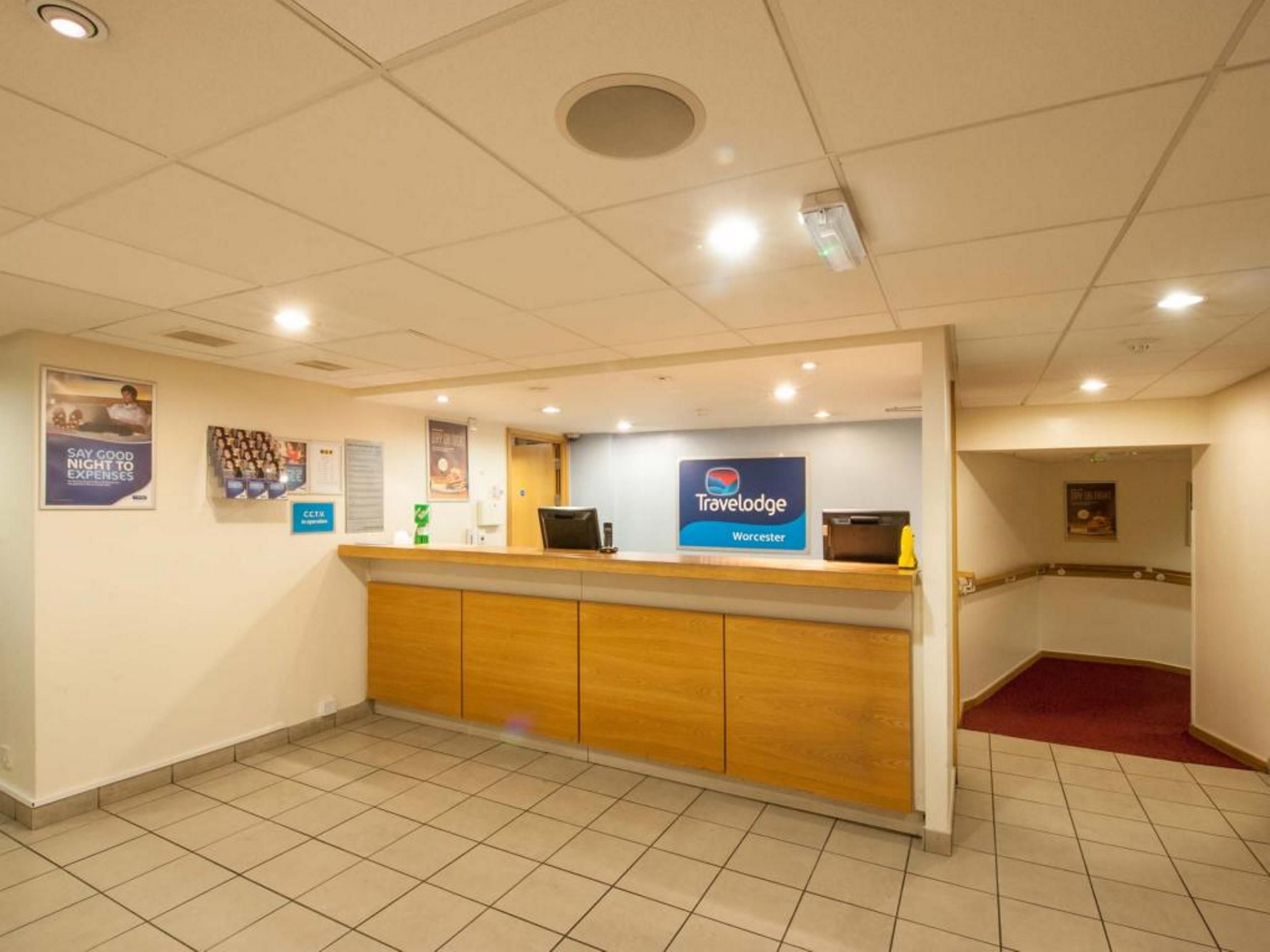 Un comptoir d’accueil avec une enseigne indiquant « Travelodge Worcester », un sol carrelé et une zone recouverte d’une moquette rouge.
