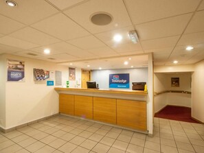 A reception desk with a sign for Travelodge Worcester, a tiled floor, and a red carpeted area. - Travelodge Worcester (Worcester)