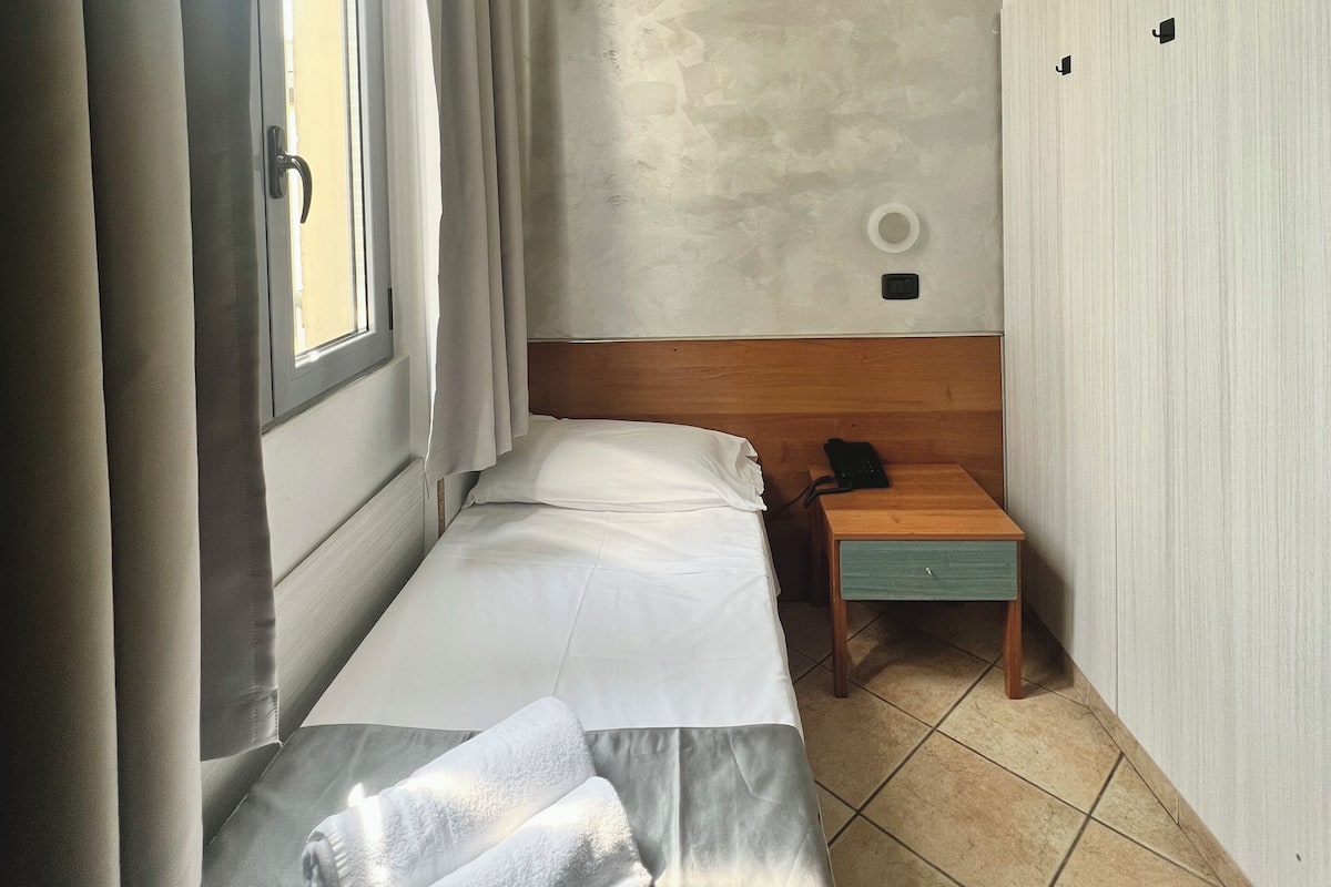 Une petite chambre avec un lit simple, une tête de lit en bois, une table de chevet avec un téléphone et une fenêtre avec des rideaux.