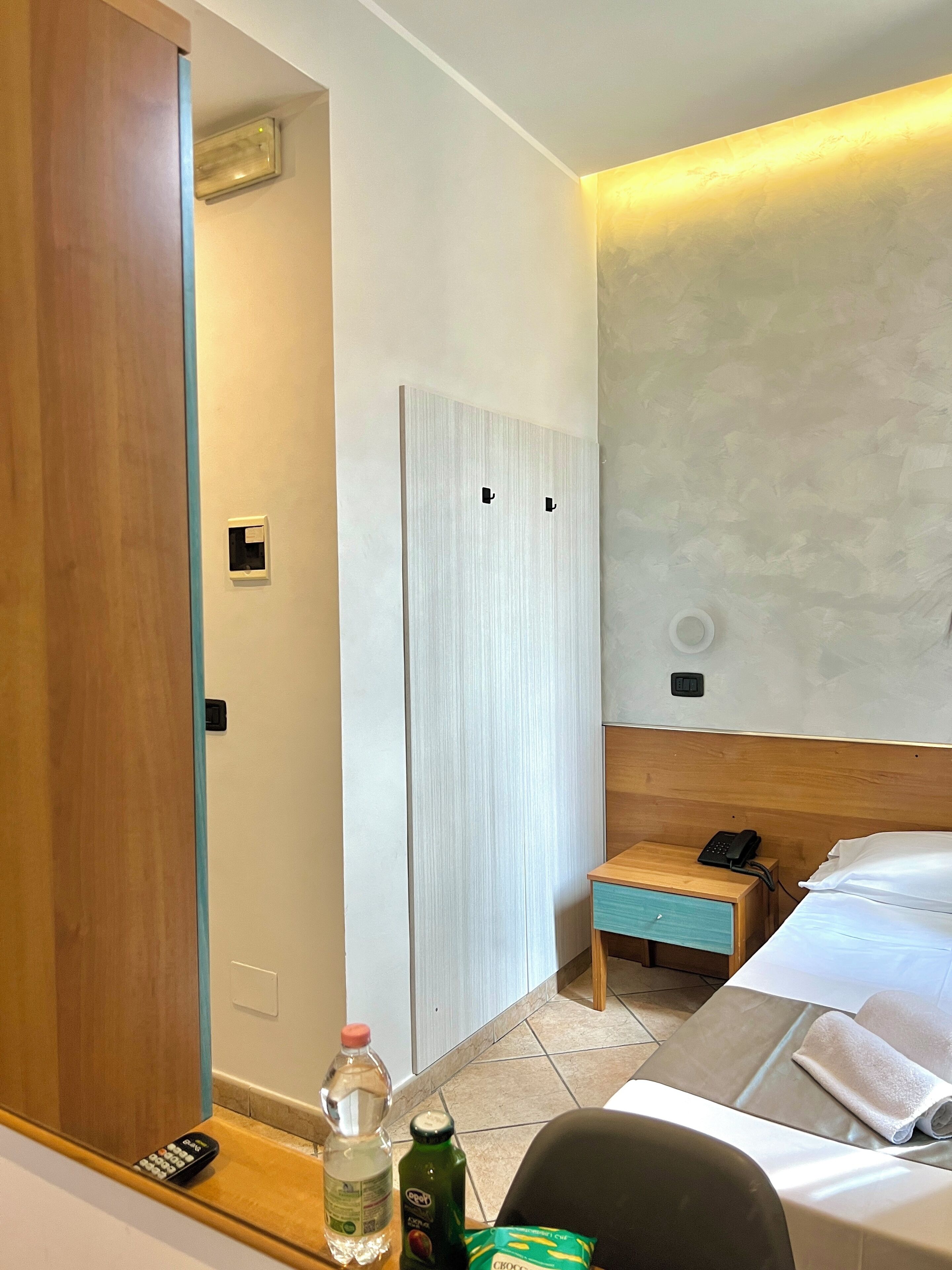 A hotel room with a wooden door, a bedside table with a remote control, a bottle of water, a green bottle, and a towel on the bed.