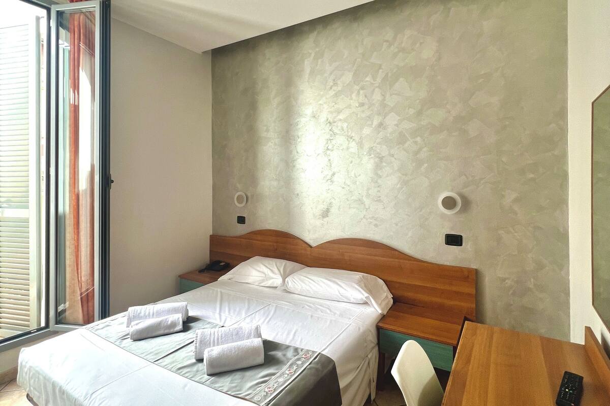 Une chambre d’hôtel avec un lit, une table de chevet en bois, un bureau avec une télécommande et une fenêtre avec des stores.