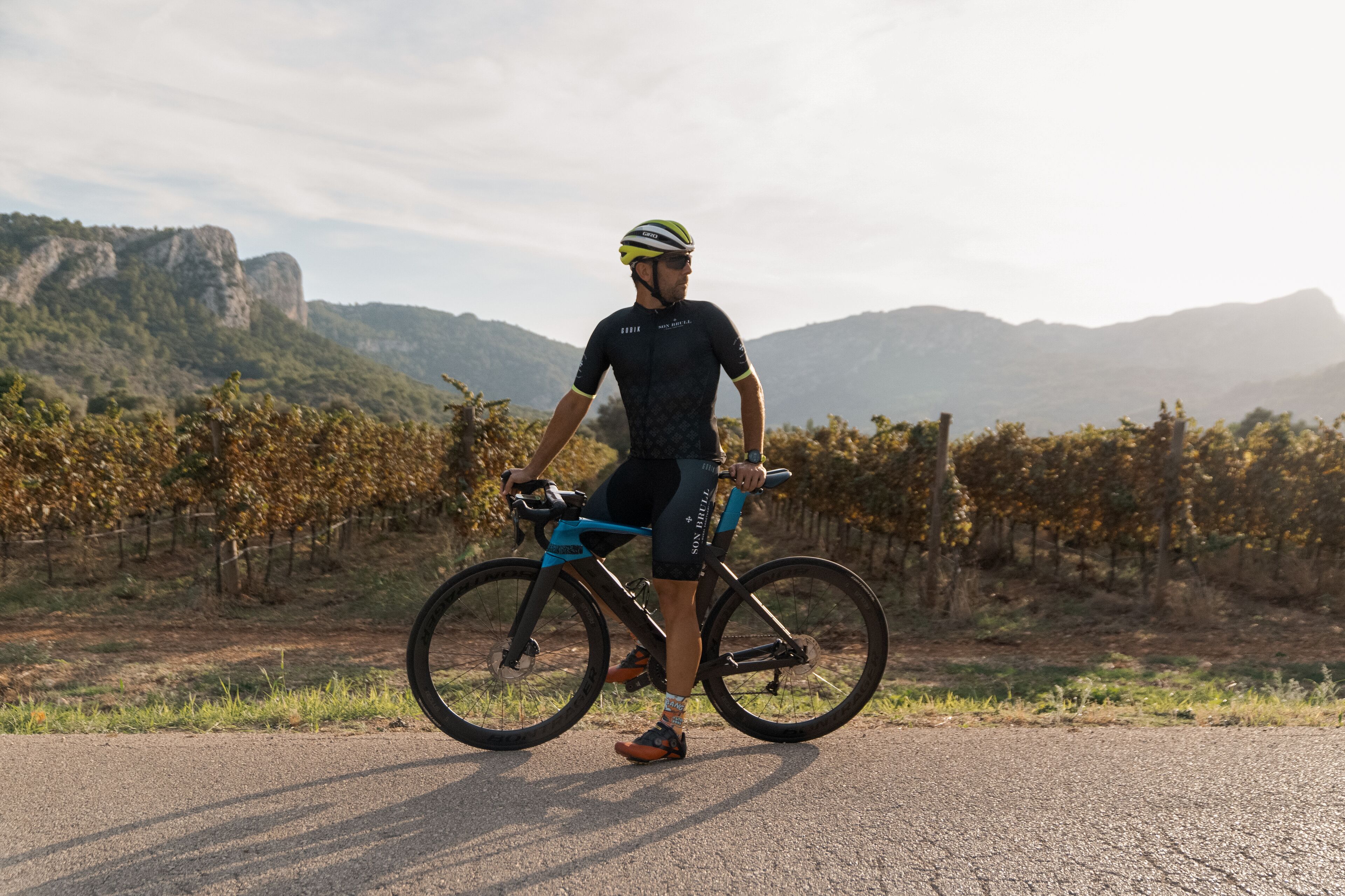 Una persona de pie junto a una bicicleta en una carretera pavimentada, con un paisaje montañoso al fondo.