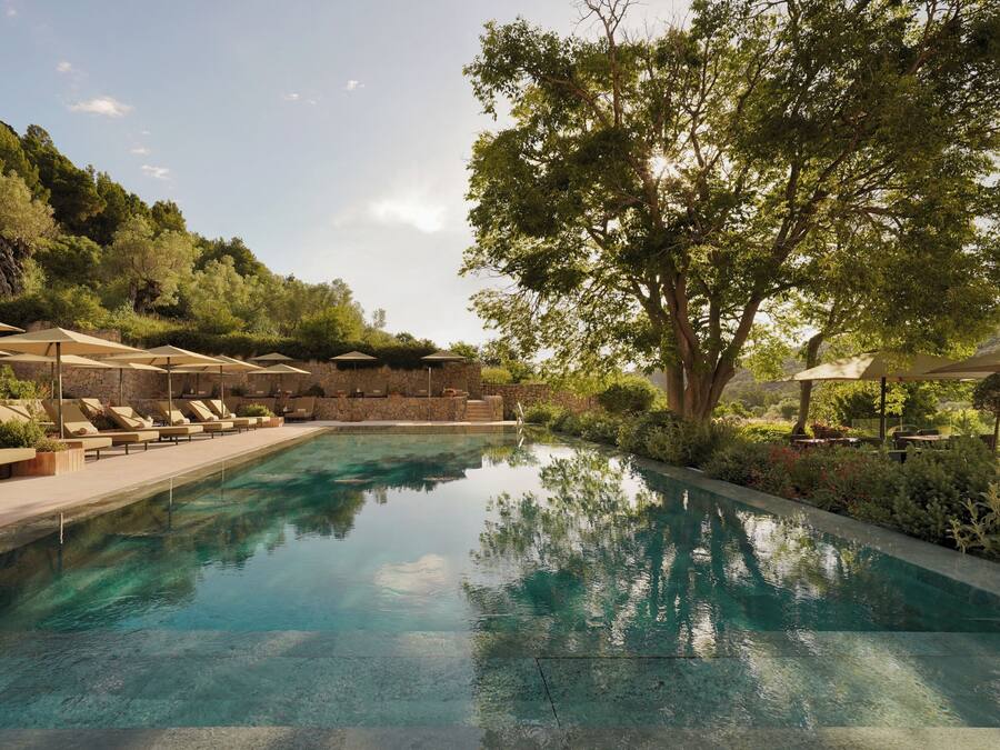 A pool with lounge chairs and umbrellas, surrounded by greenery and a stone wall.