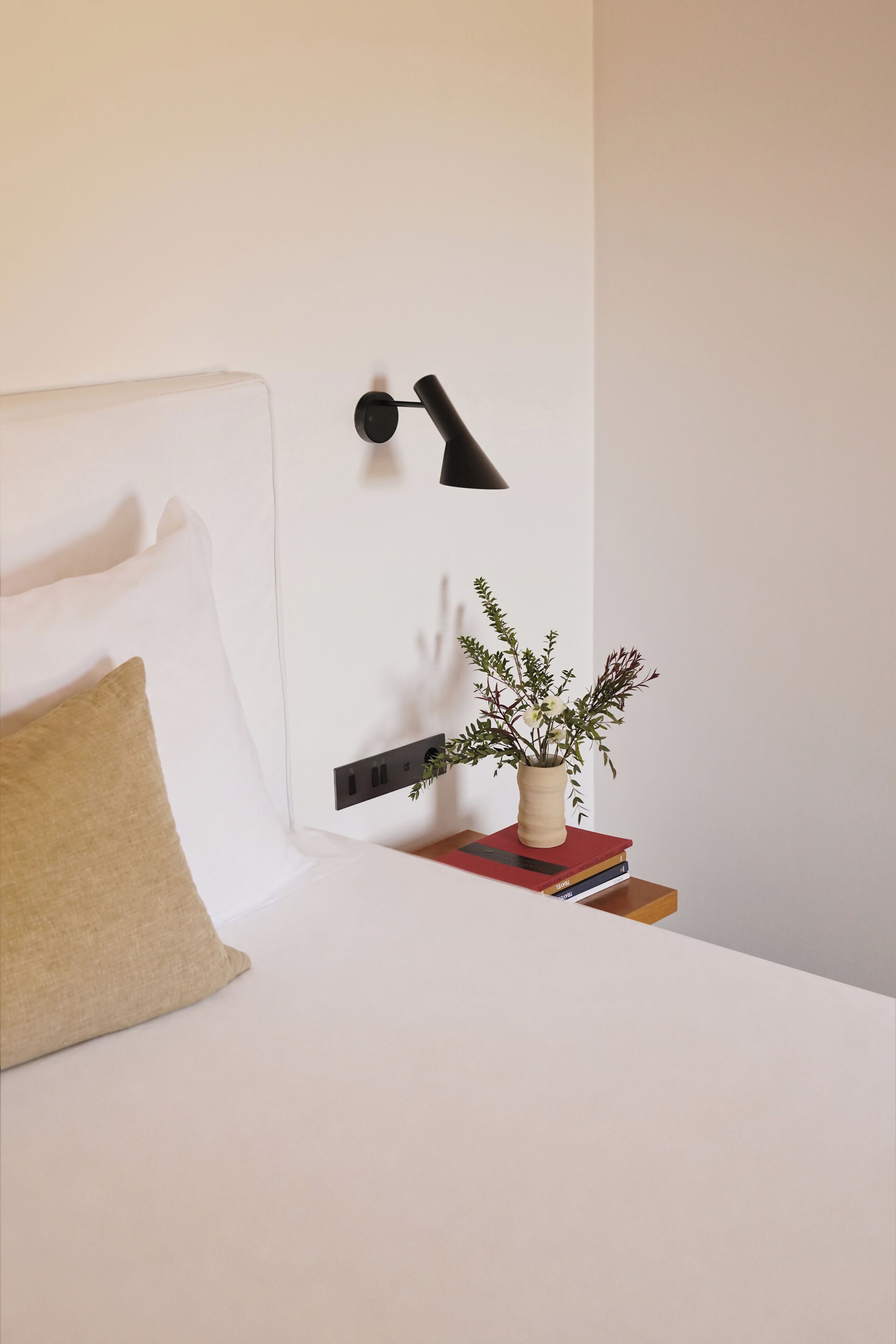 Una cama bien tendida con una sola almohada, una lámpara de noche y una pequeña planta en maceta sobre una mesita de noche.