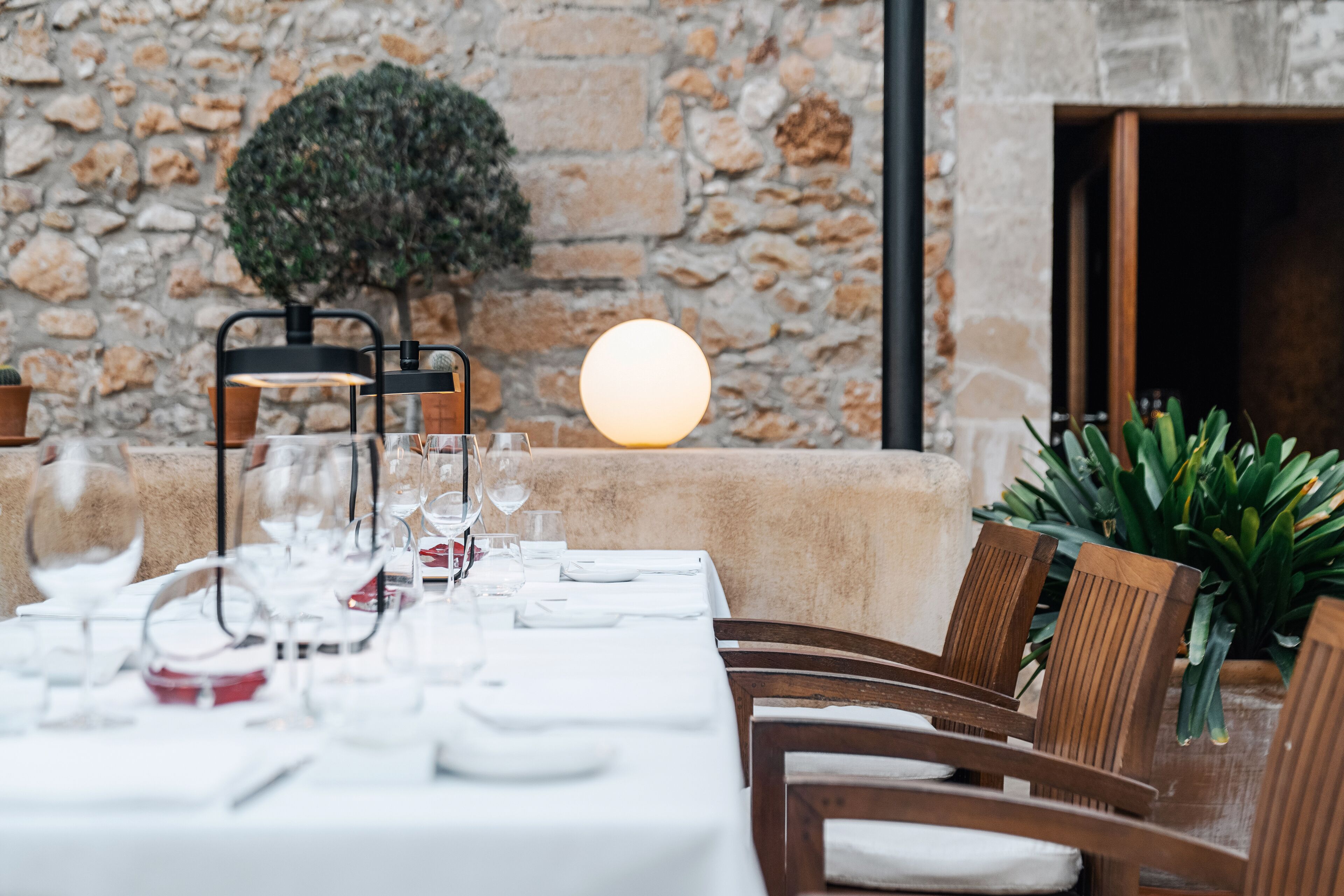 Mesa de comedor vestida con mantel blanco, copas y platos, con una pared de piedra y un pequeño árbol al fondo.
