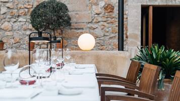 Mesa de comedor vestida con mantel blanco, copas y platos, con una pared de piedra y un pequeño árbol al fondo.