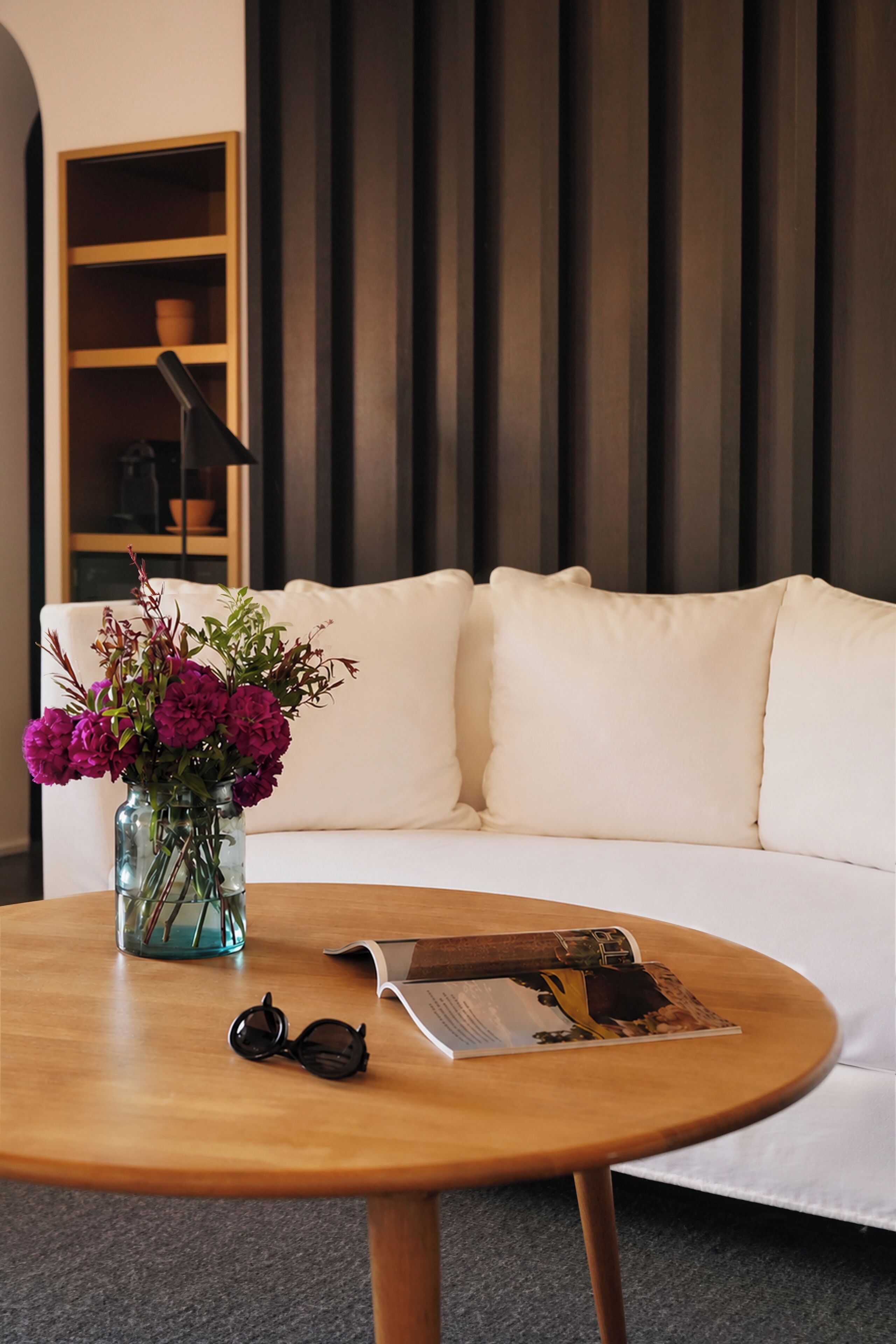 Un salon moderne avec un canapé blanc, une table basse ronde en bois, un vase avec des fleurs, une paire de lunettes et un magazine.