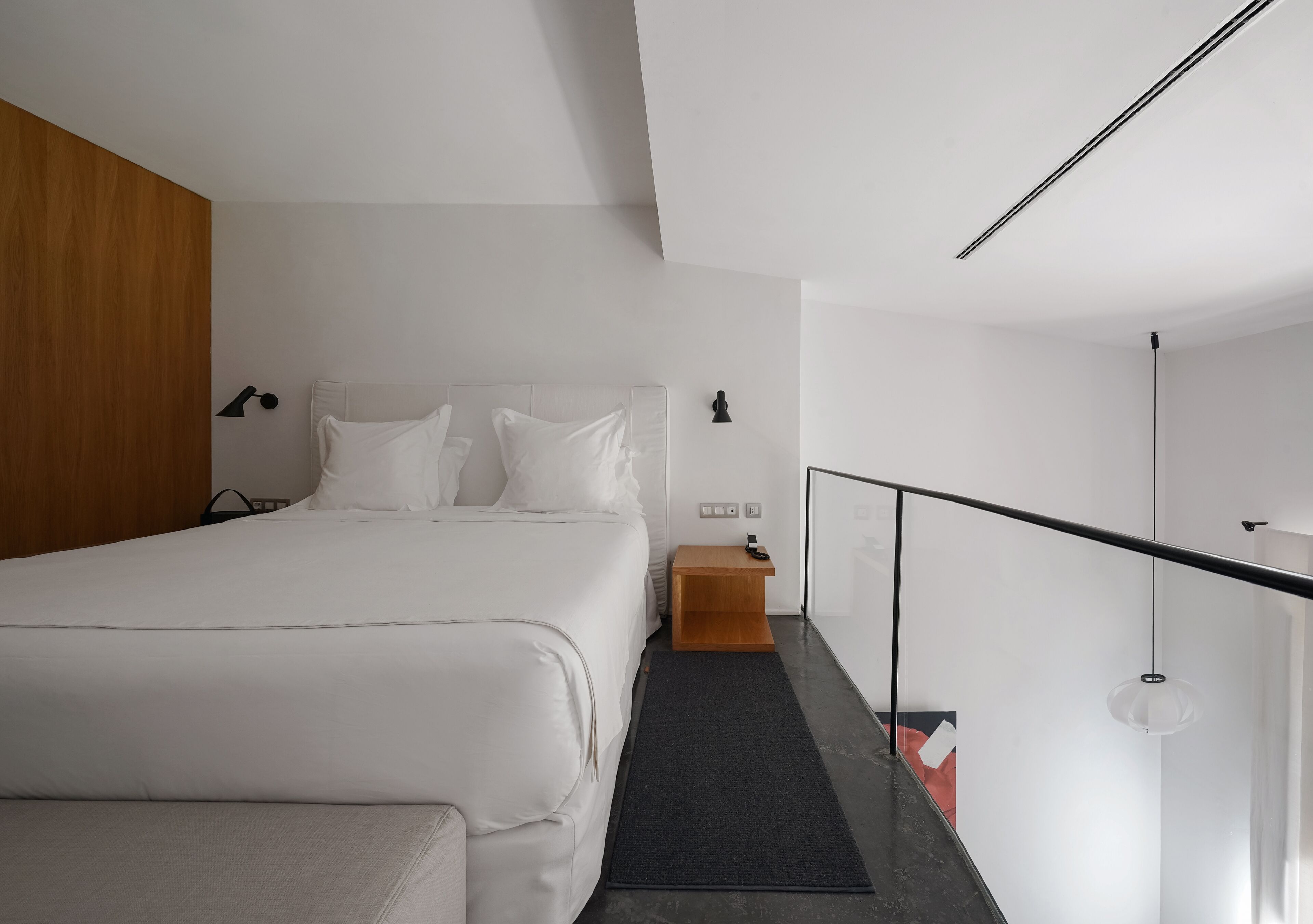 Een moderne hotelkamer met een groot bed, een houten hoofdbord, een klein houten nachtkastje, een zwart vloerkleed en een glazen balustrade.