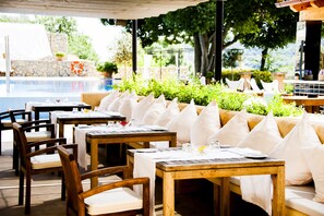 An outdoor dining area with wooden tables and chairs, a pool in the background, and a stone wall with greenery. - Son Brull Hotel & Spa (Pollenca)