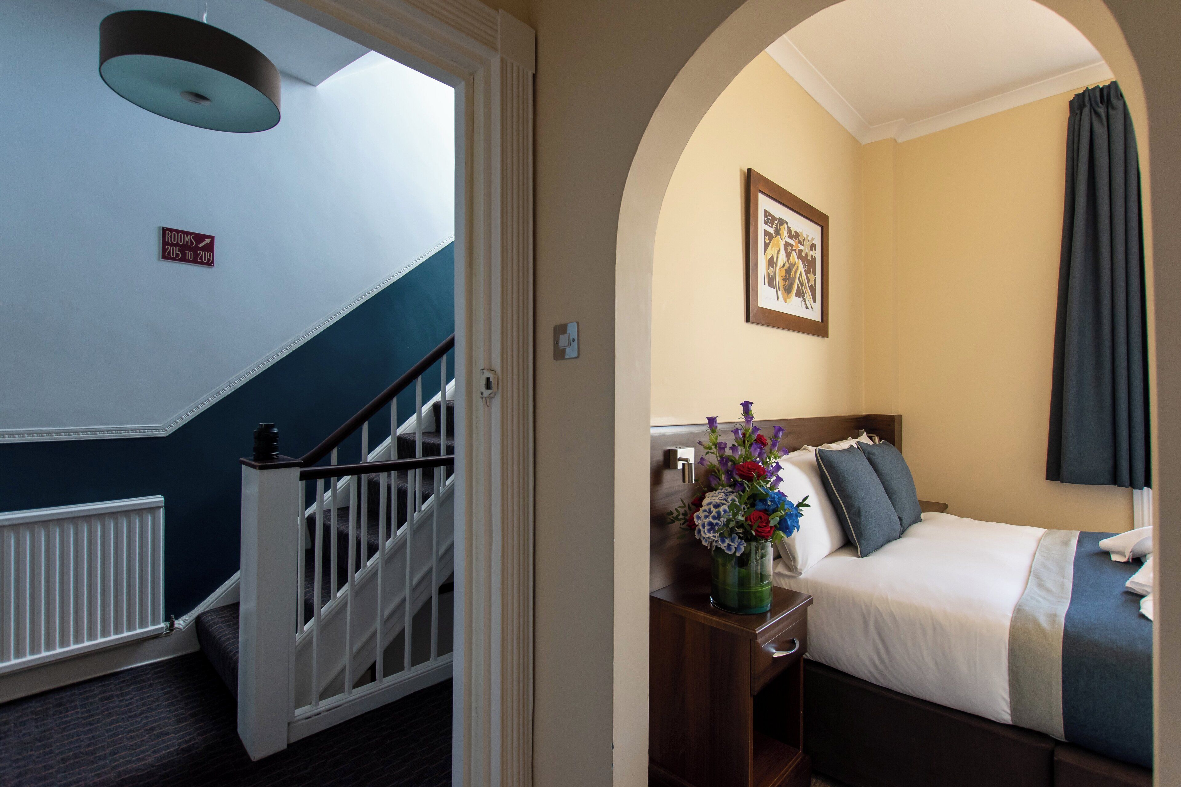 Ein Hotelzimmer mit einem Bett, einem Nachttisch, einem Blumenstrauß, einem gerahmten Bild an der Wand und einer Treppe, die in ein weiteres Zimmer führt.