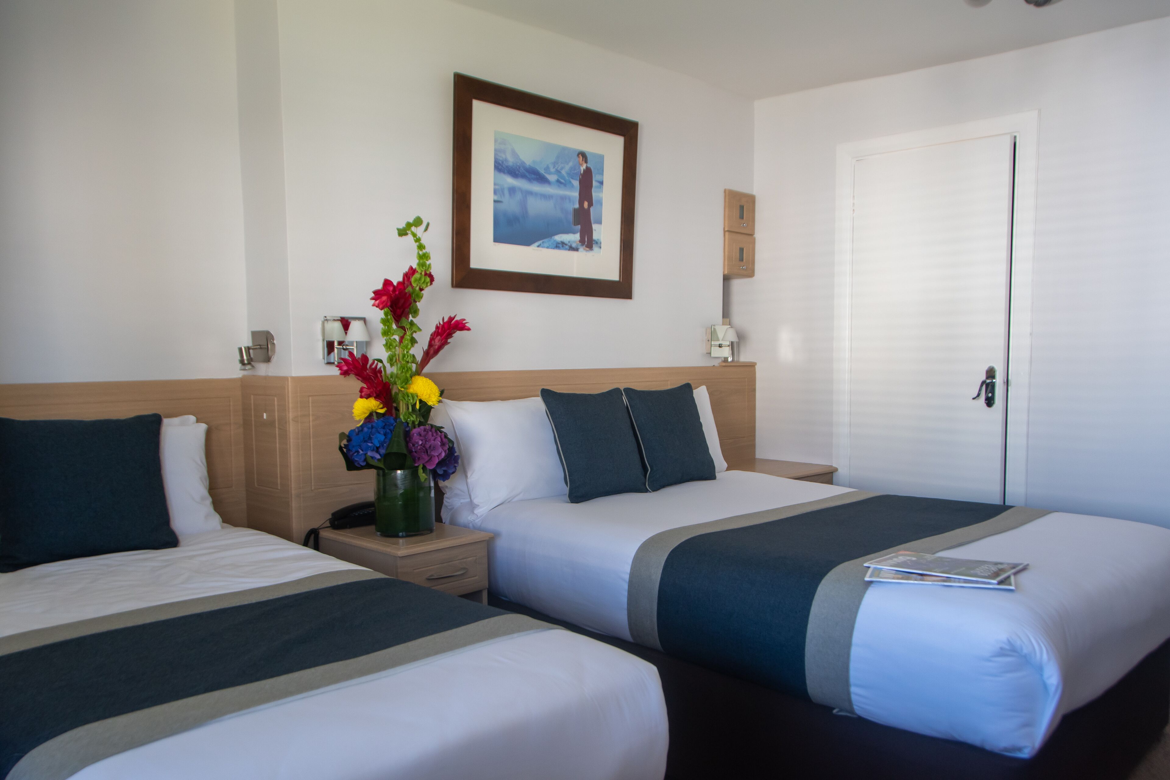 A hotel room with two beds, a nightstand, a vase with flowers, a framed picture on the wall, and a door.