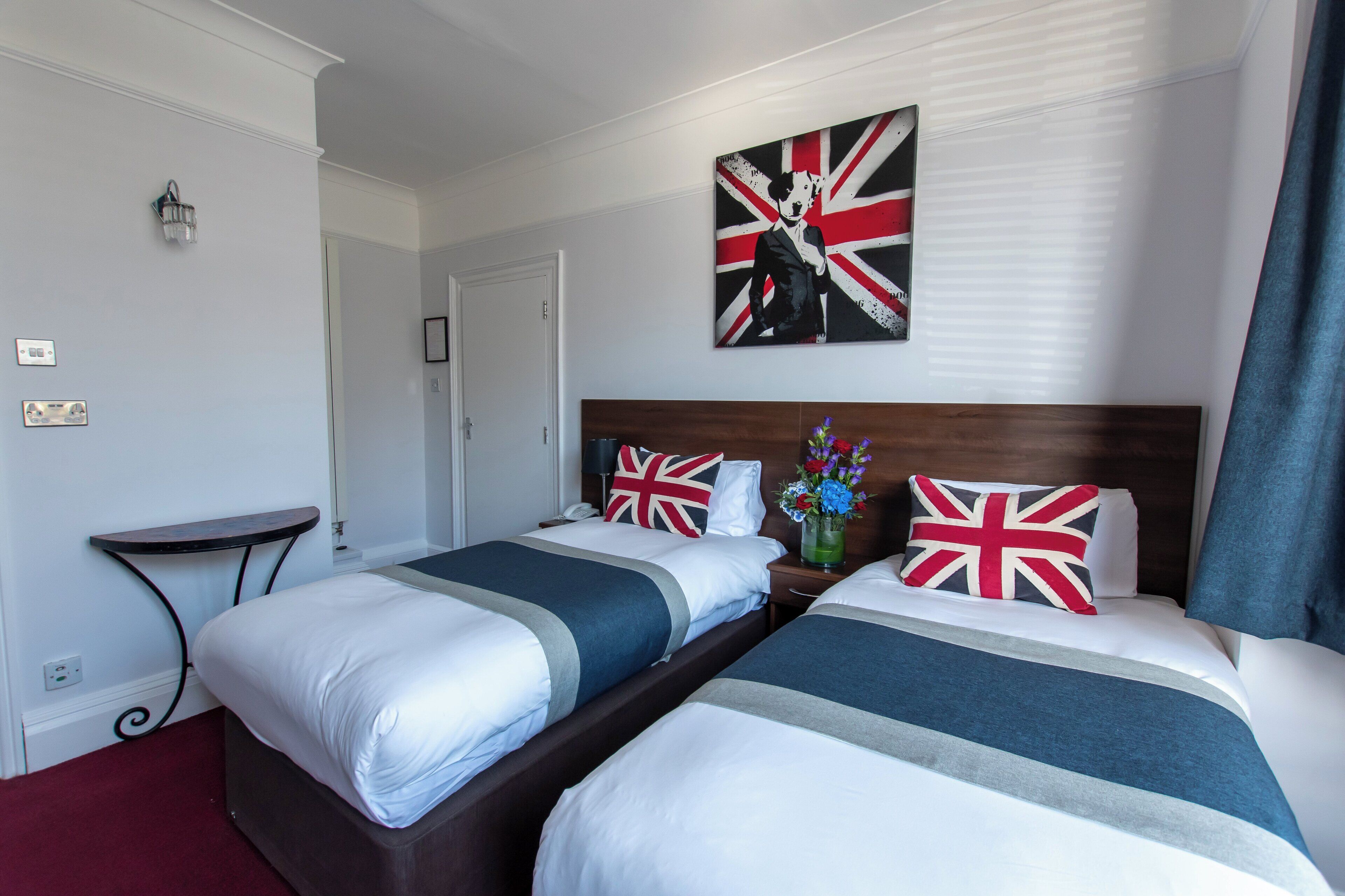 Une chambre d’hôtel avec deux lits, une table de chevet, une table et un tableau représentant une figure tenant un drapeau Union Jack.