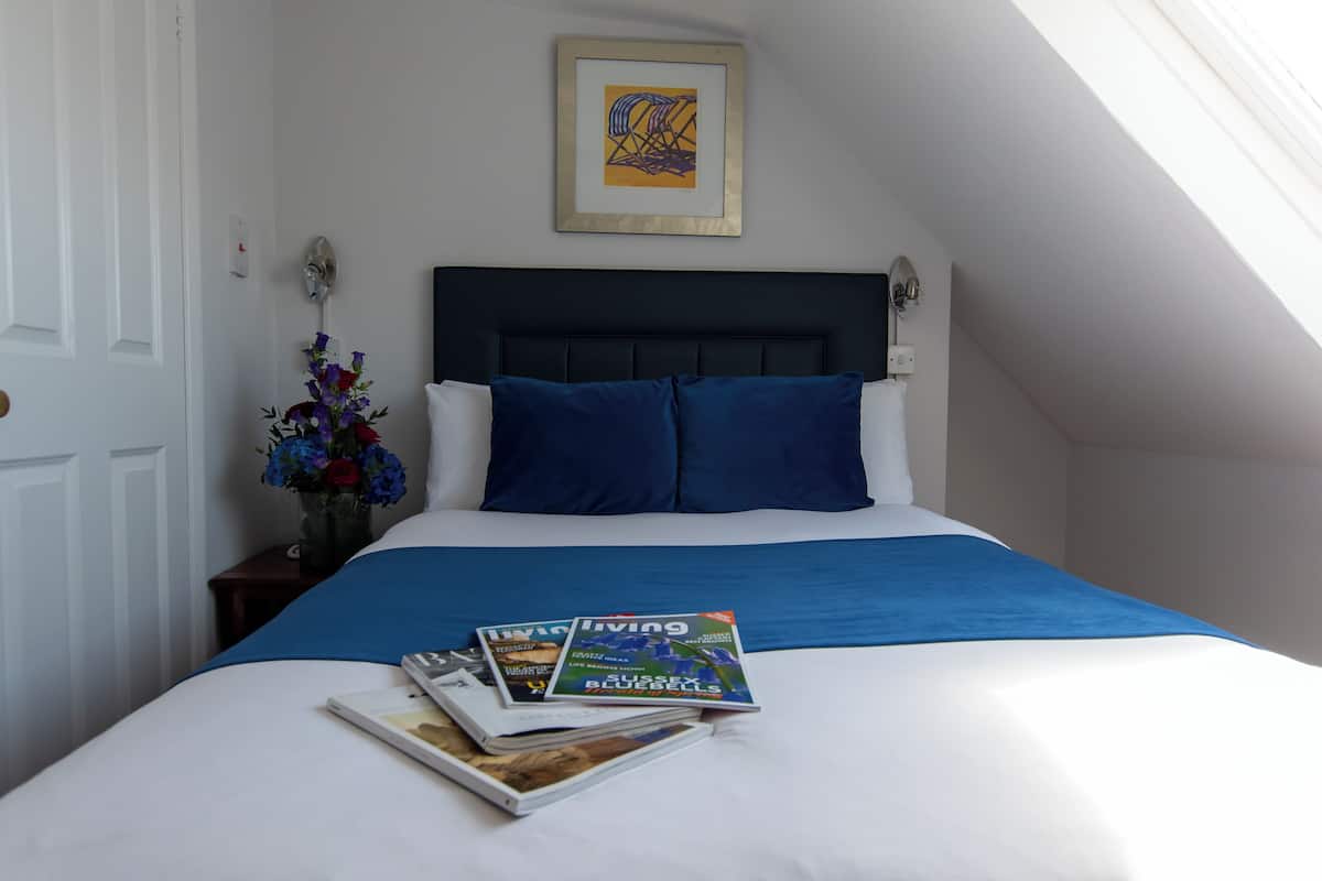 A neatly made bed with a blue bedspread and pillows, a framed abstract painting above it, and a vase with flowers on a nightstand beside the bed.