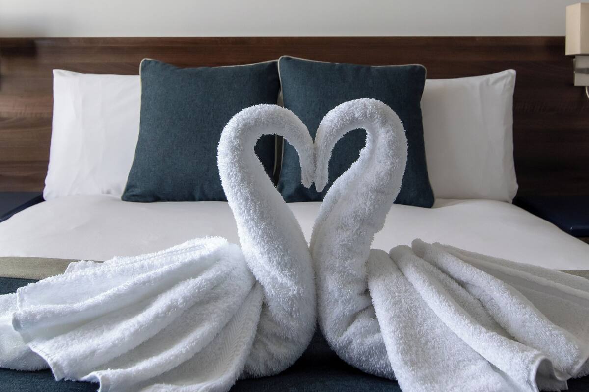 A neatly made bed with two towels arranged in the shape of swans, a blue pillow, and a painting of a person on a boat in the background.
