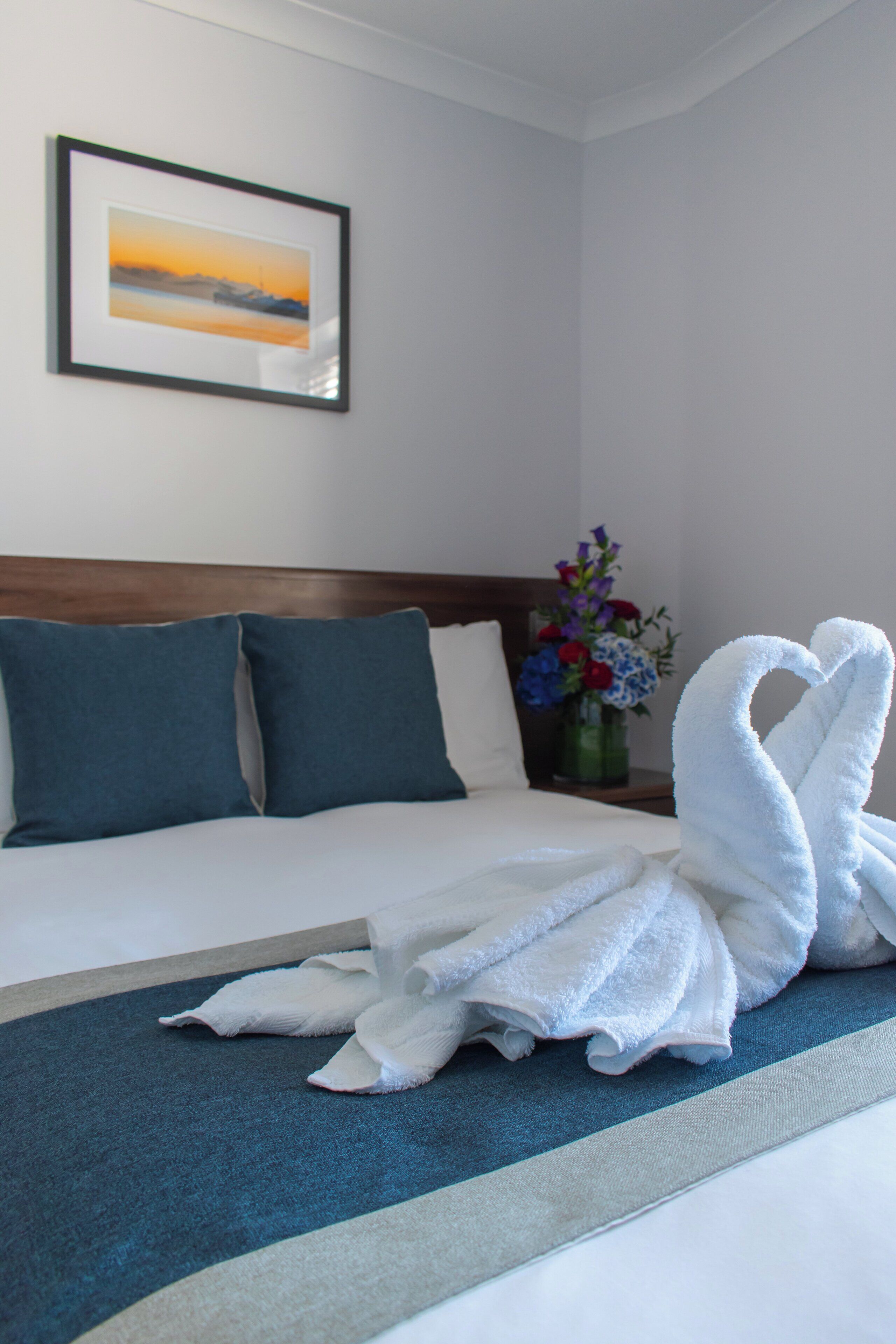 A hotel room with a neatly made bed, a framed picture on the wall, and a vase of flowers on the nightstand.