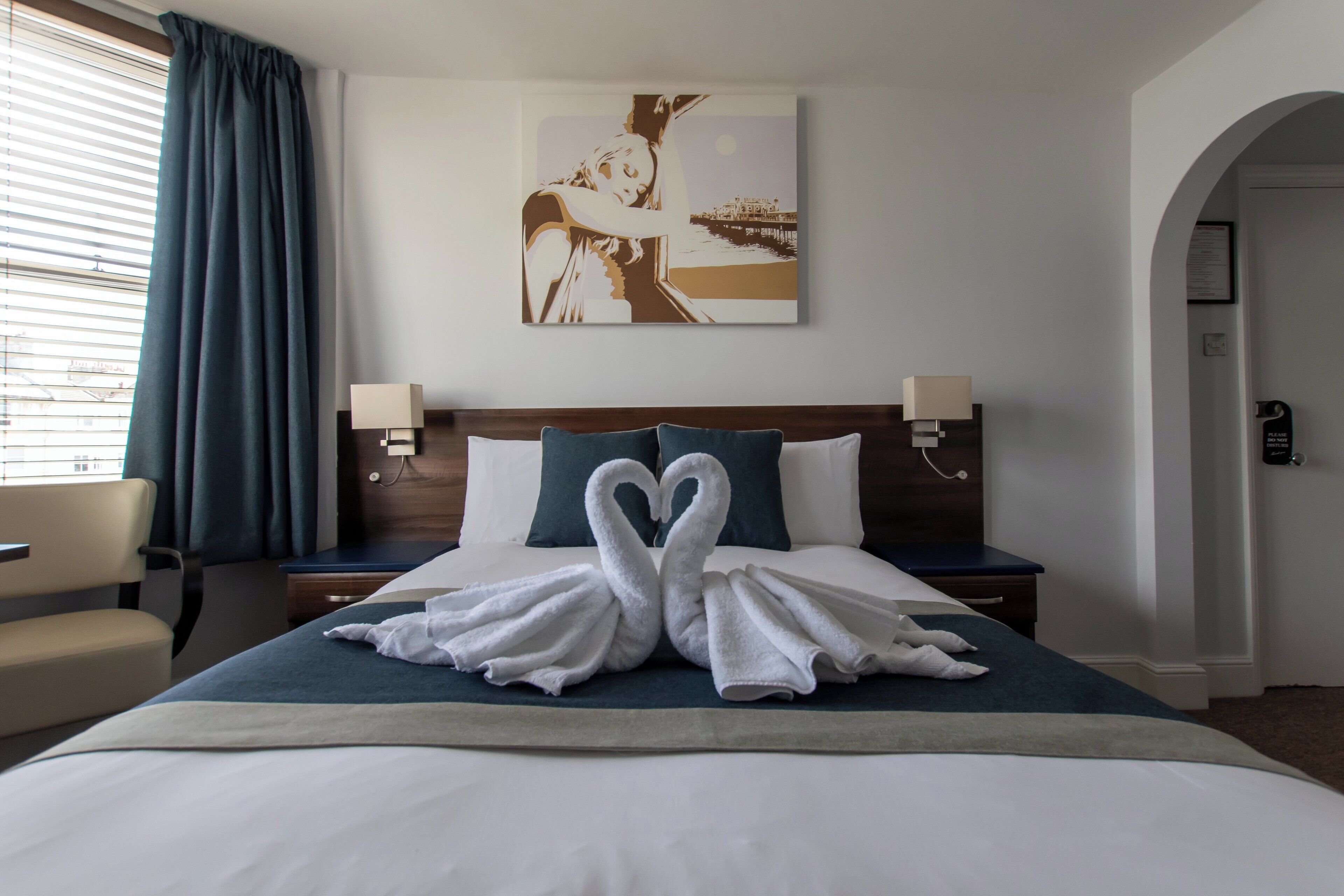 Habitación de hotel con cama, dos mesitas de noche, lámpara, silla, un cuadro en la pared y vistas al exterior a través de la ventana.