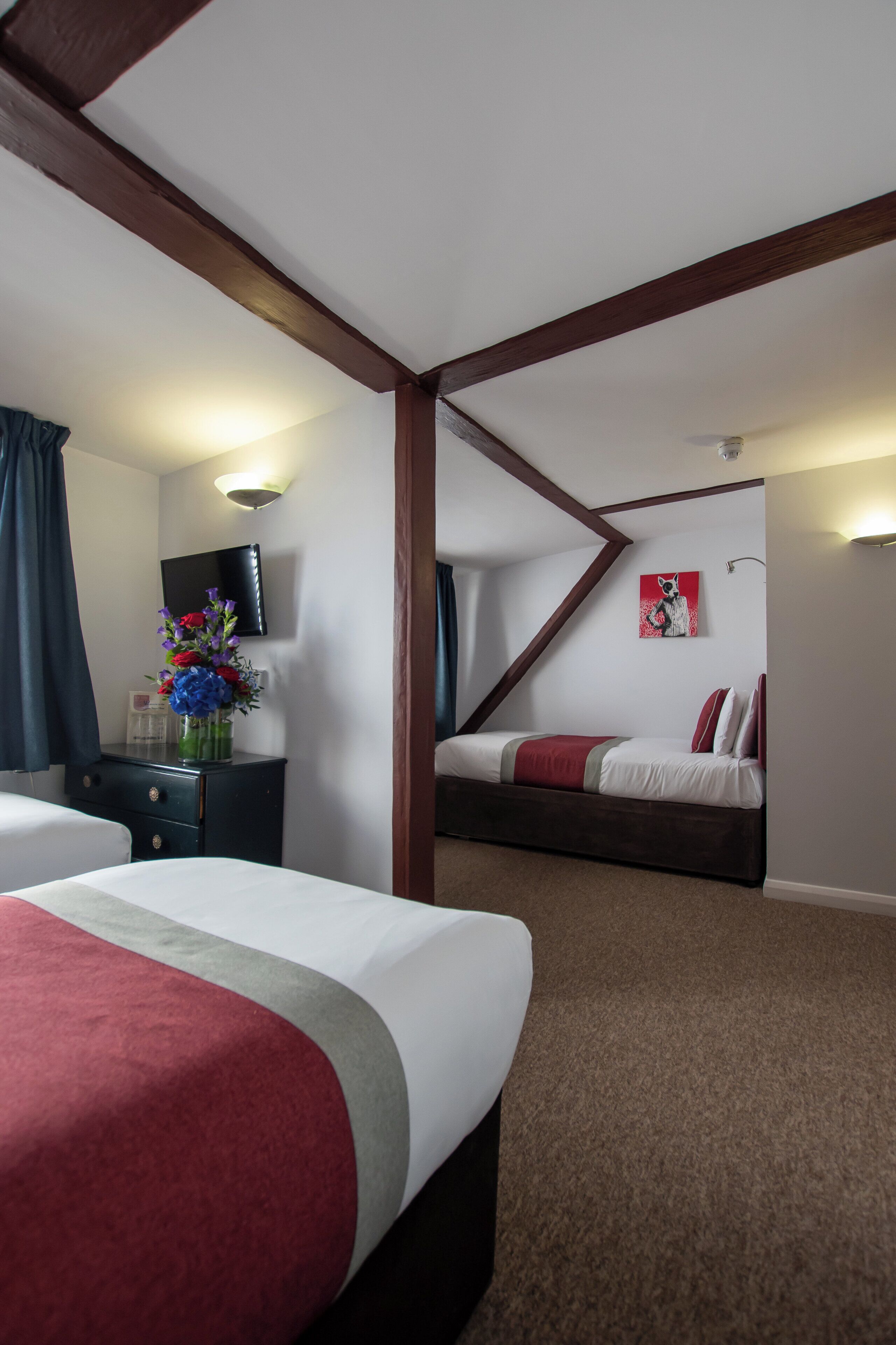 Une chambre d’hôtel avec deux lits, une télévision, un bureau agrémenté d’un vase de fleurs et un tableau au mur.