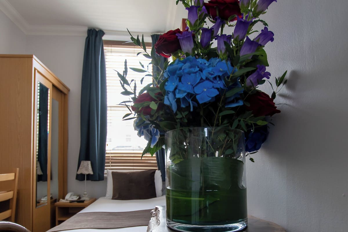 A hotel room with a bed, a wooden wardrobe, a desk with a magazine, and a vase with flowers.