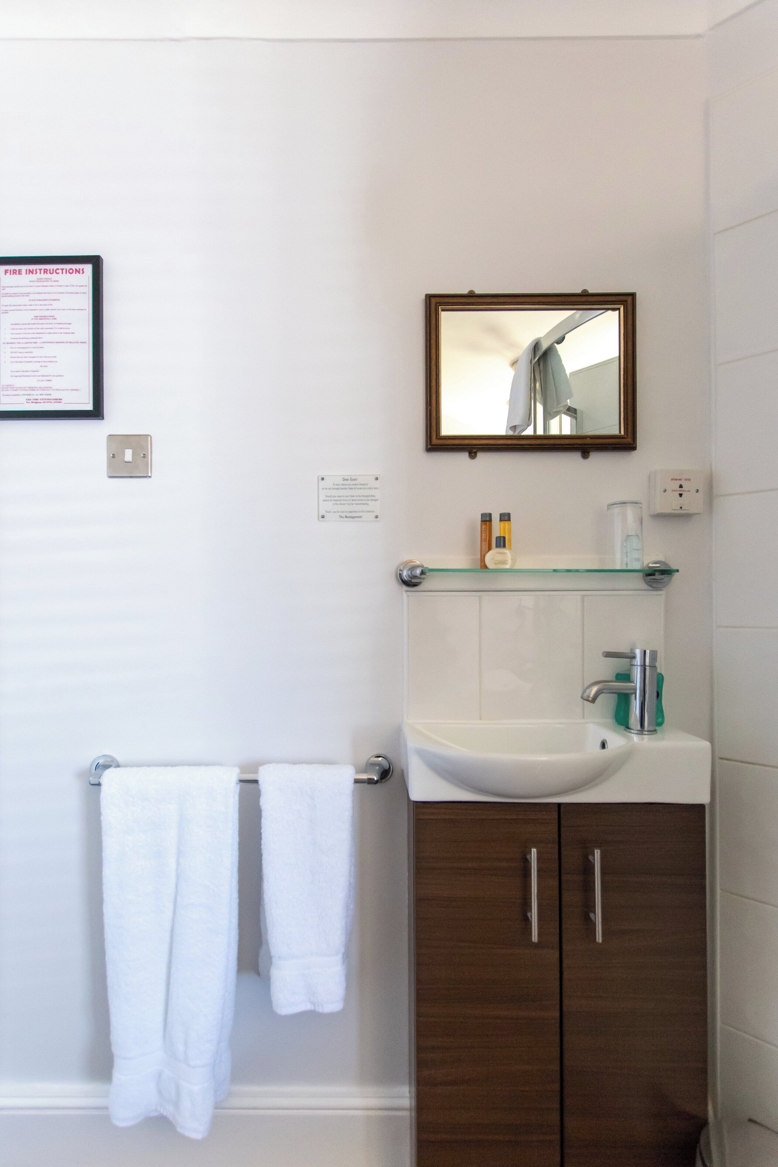 A bathroom with a white sink, wooden cabinets, a glass shelf with toiletries, and a framed mirror.