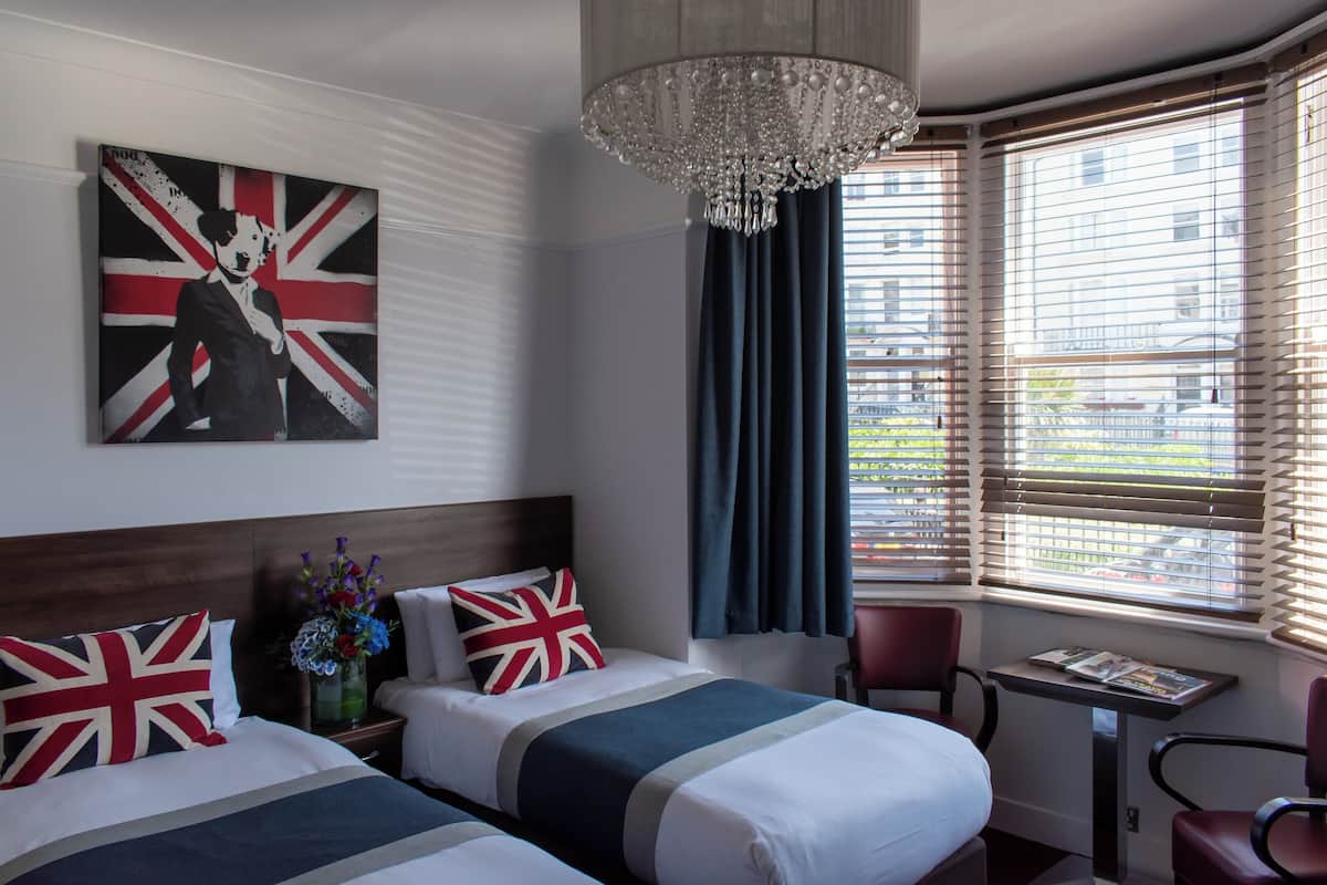A hotel room with two beds, a chandelier, a table with magazines, and a window with blinds.