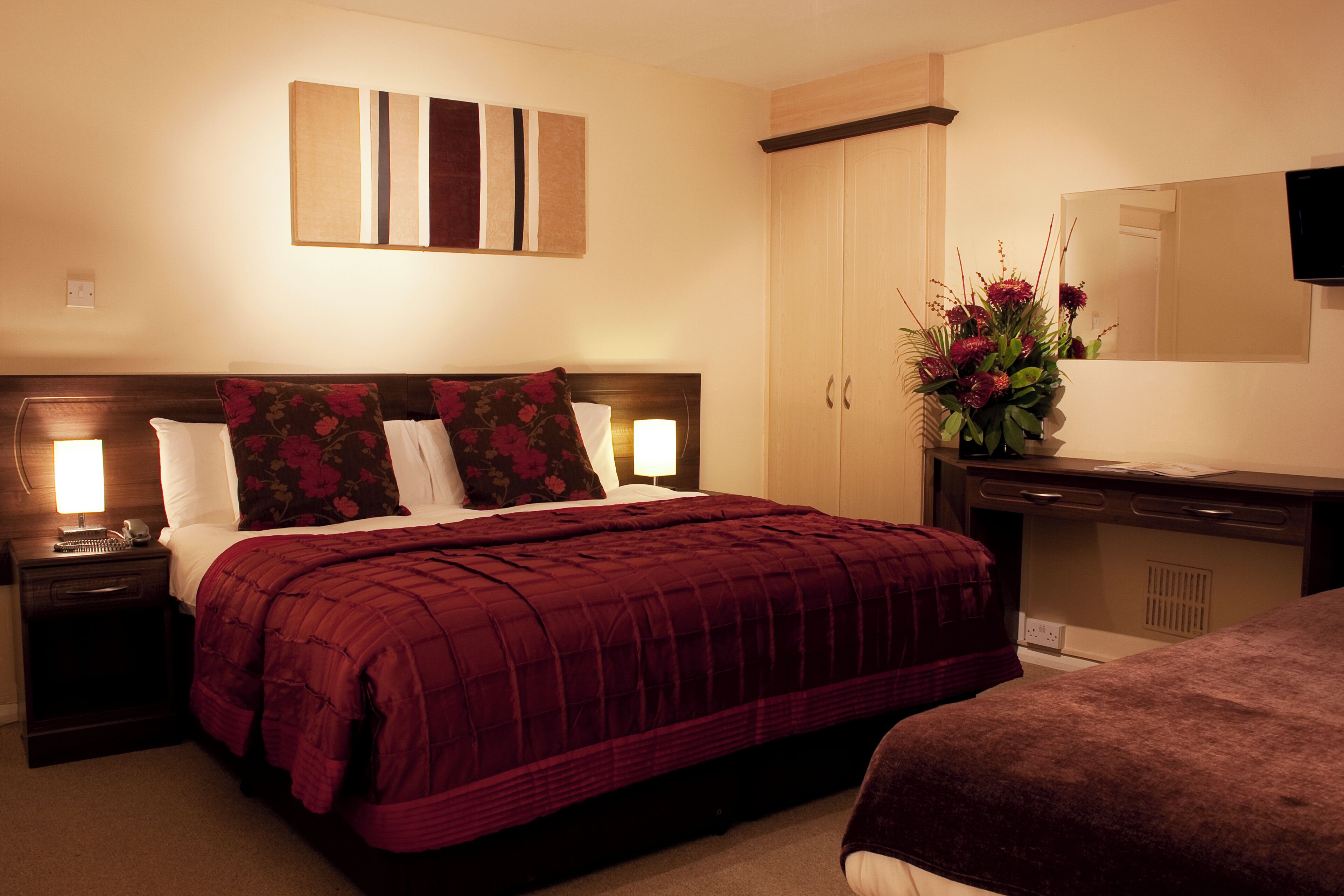 A hotel room with a large bed, a nightstand, a dresser, a mirror, and a vase with flowers.