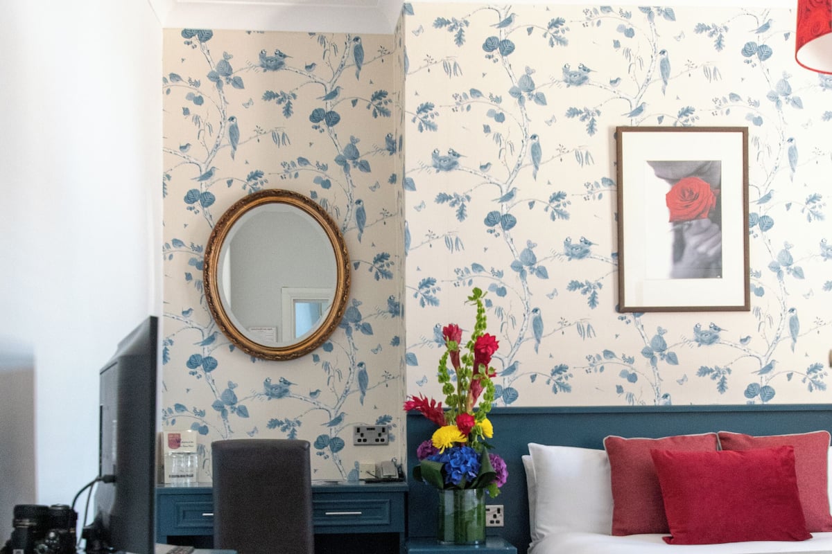 A hotel room with a bed, a desk, a chair, a mirror, a vase with flowers, and a framed picture on the wall.